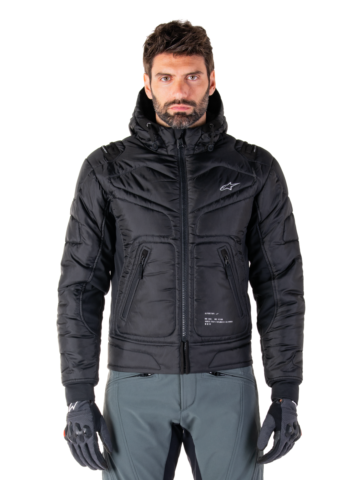 Alpinestars Mohobbs vandtæt jakke, sort polstret bomber-motorcykeljakke, quiltet poly-materiale med lynlås foran og lynlåslommer, lille hvidt logo på brystet, båret af en model med grå bukser og handsker, urban streetwear-design til bykørsel