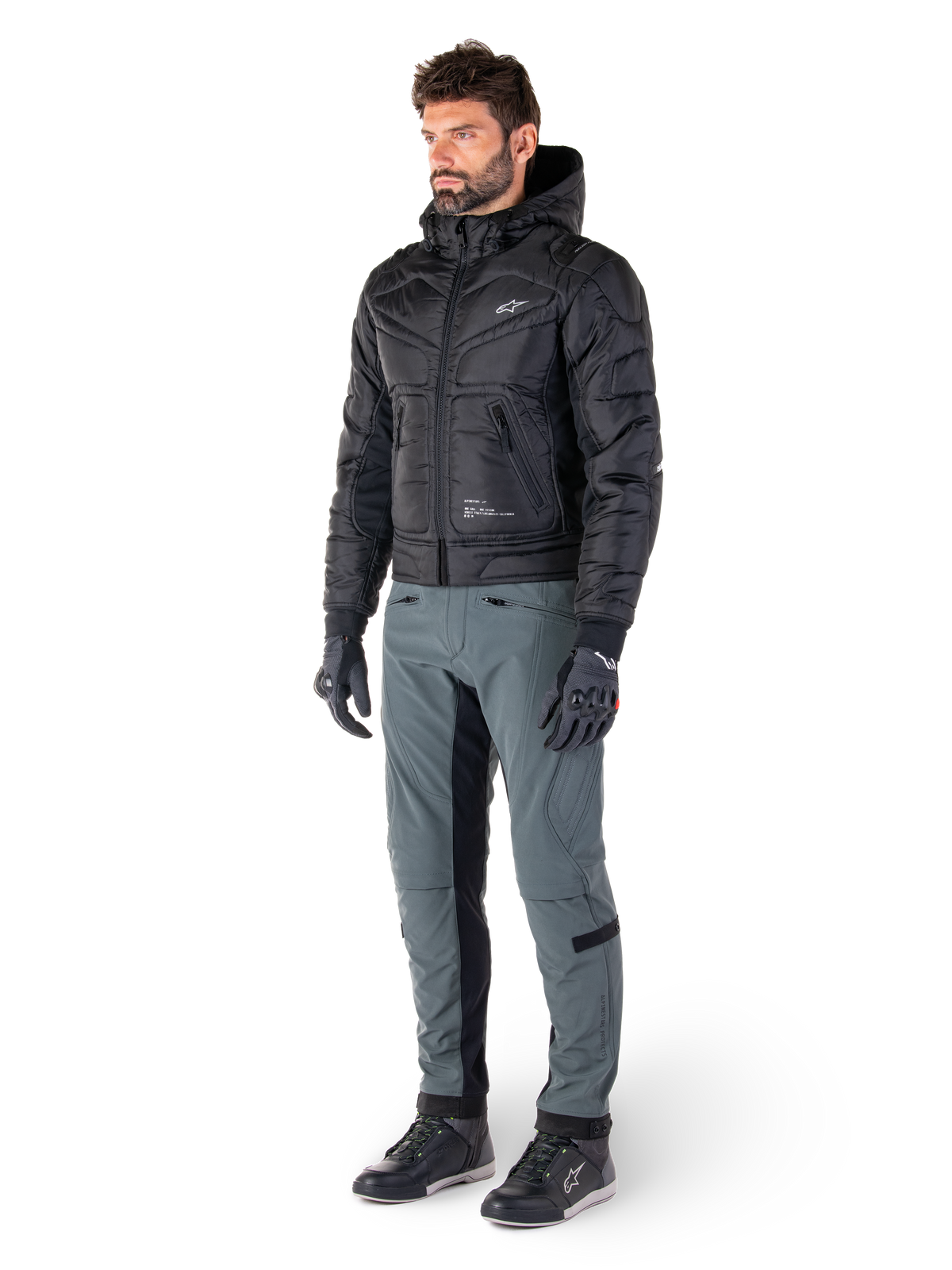 Alpinestars Mohobbs Waterproof Jacket, sort, quiltet urban motorcykel-bomberjakke med hætte, båret af en model med grå kørebukser og sorte handsker, med hvidt brystlogo, moderne streetwear-inspireret beskyttelsesudstyr