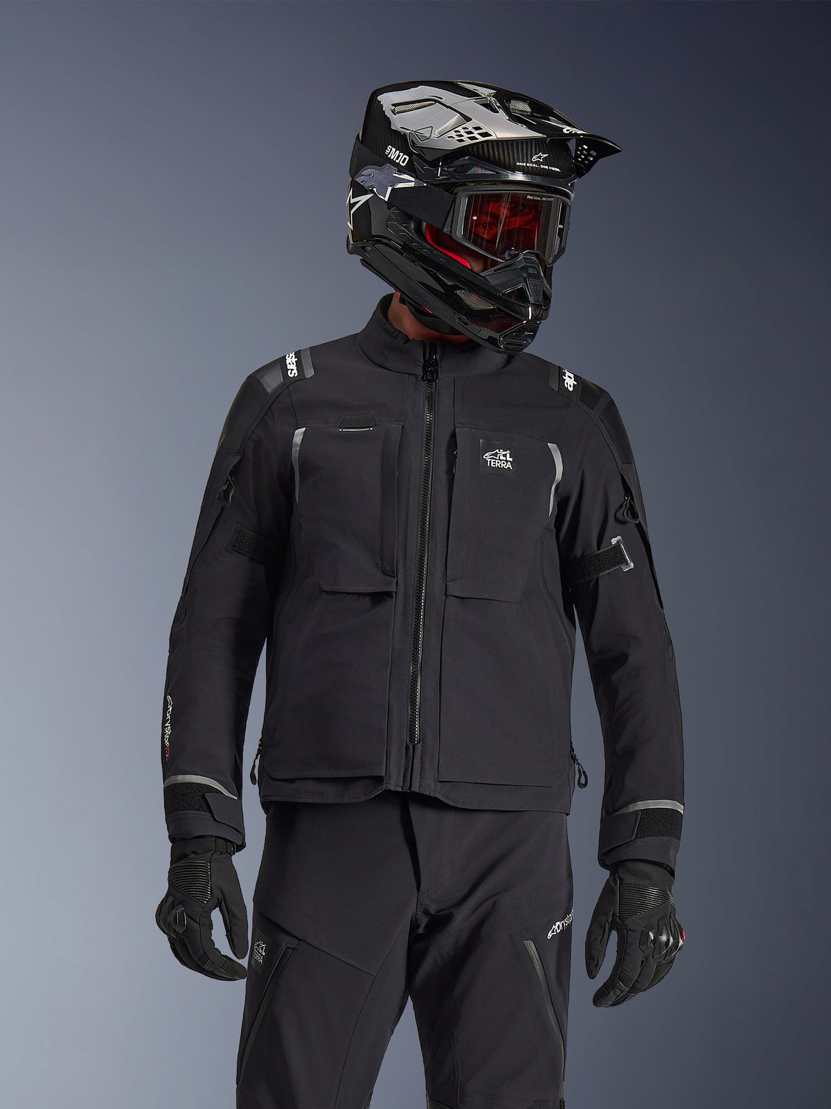 Adventure rider wearing the Alpinestars Andes Pro Drystar® XF Jacket in Black and Dark Gray, featuring All Terra chest branding, paired with matching technical pants and a black adventure helmet with goggles, professional touring gear set.