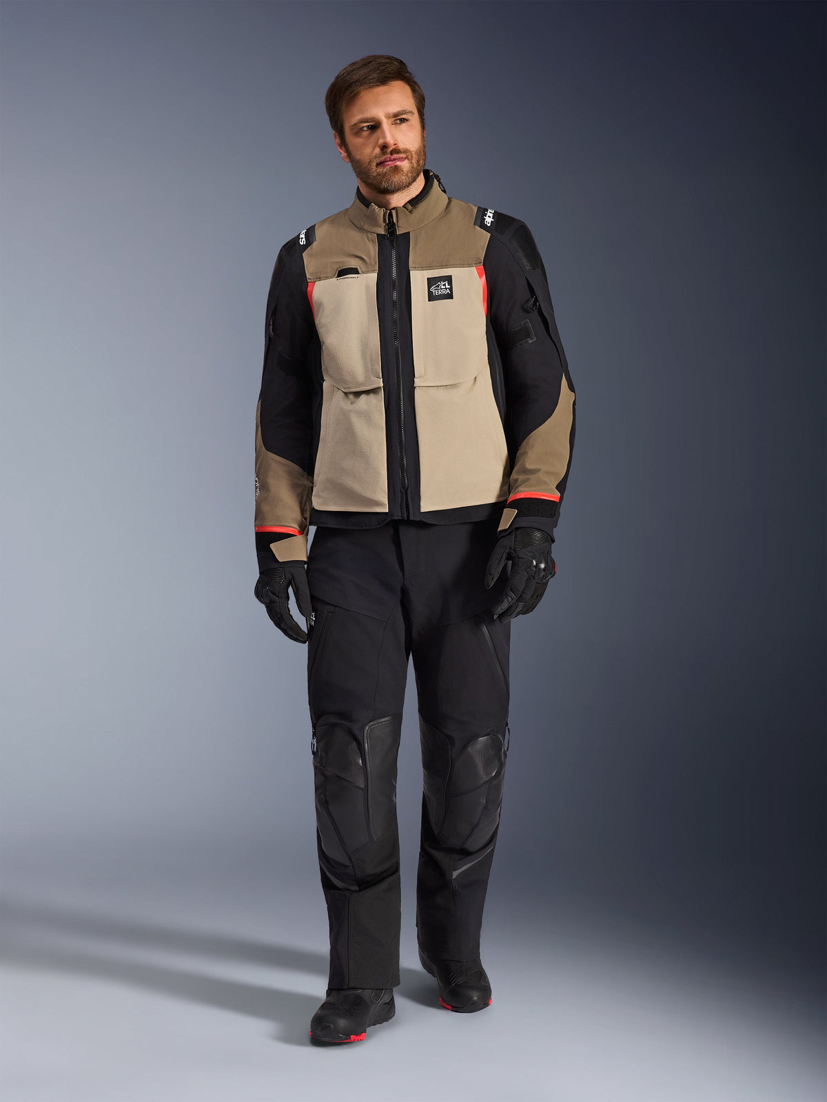 Mandlig model iført Alpinestars Andes Pro Drystar® XF-jakke i Walnut Aluminum, tan- og sort adventure-touringjakke med røde detaljer, kombineret med sorte motorcykelbukser, handsker og støvler, studiefoto i fuld figur af beskyttende køreudstyr.