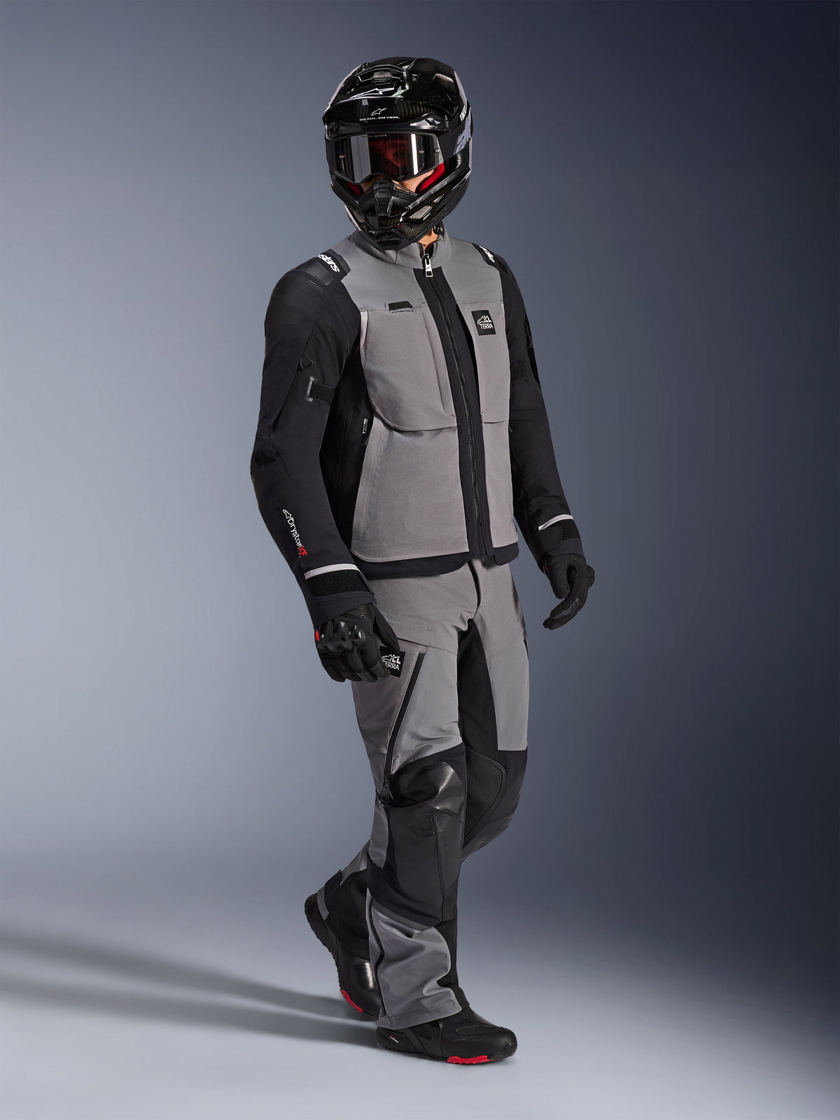 Adventure-rytter iført Alpinestars Andes Pro Drystar® XF-jakke i mørkegrå og sort, matchende grå og sorte adventure-bukser, sort full-face hjelm med goggles og sorte motorcykelhandsker, studiebillede af professionelt udstyr til langdistance-touring