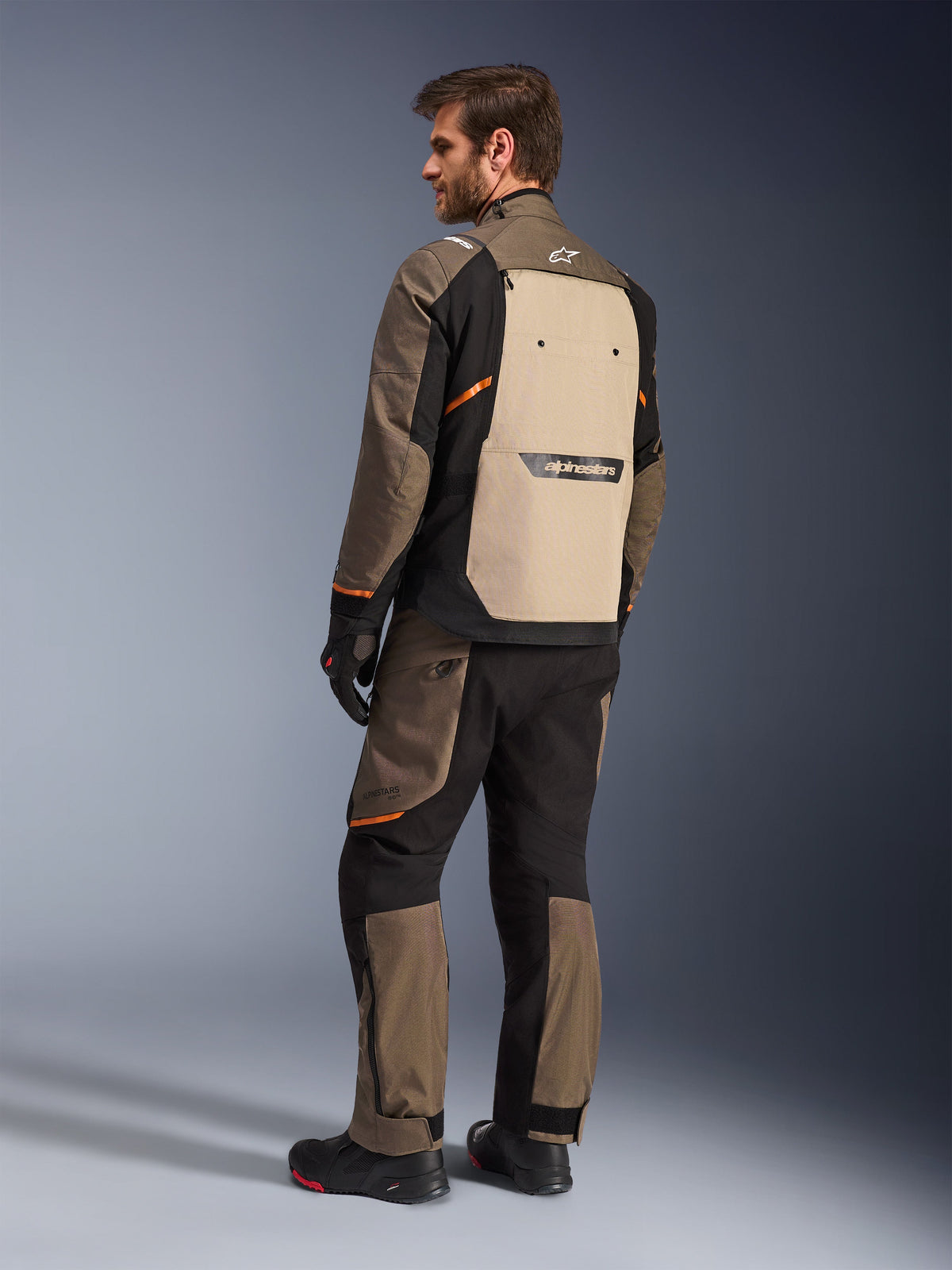 Alpinestars Cusco Drystar® Jakke i Walnut Aluminum Black, set bagfra med sandfarvet rygpanel og orange detaljer, båret med matchende motorcykelbukser, sorte handsker og støvler, professionelt adventure touring-udstyr
