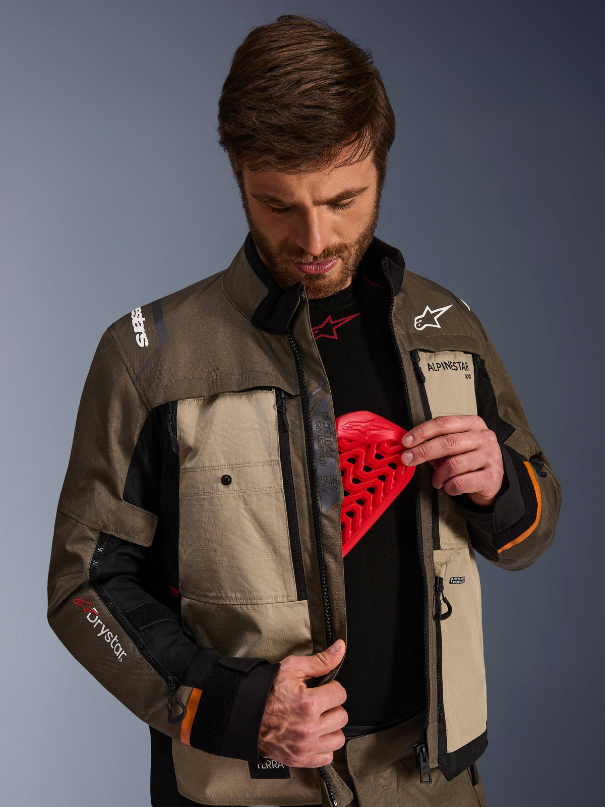 Alpinestars Cusco Drystar® jakke, Walnut Aluminum Black, mand der demonstrerer brystbeskytterlommen med en rød beskyttelsesindsats, tekstil motorcykeljakke med ventilationsåbninger med lynlås, branding til adventure touring