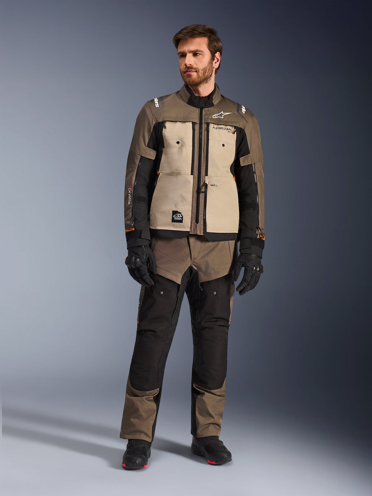 Alpinestars Cusco Drystar®-jakke i Walnut Aluminum Black, båret af en mandlig kører med matchende adventure touring-bukser, sorte handsker og støvler, med sandfarvede brystpaneler, sorte ærmer med diskrete orange detaljer og funktionelle ventilationslynlåse til motorcykelkørsel på vej.