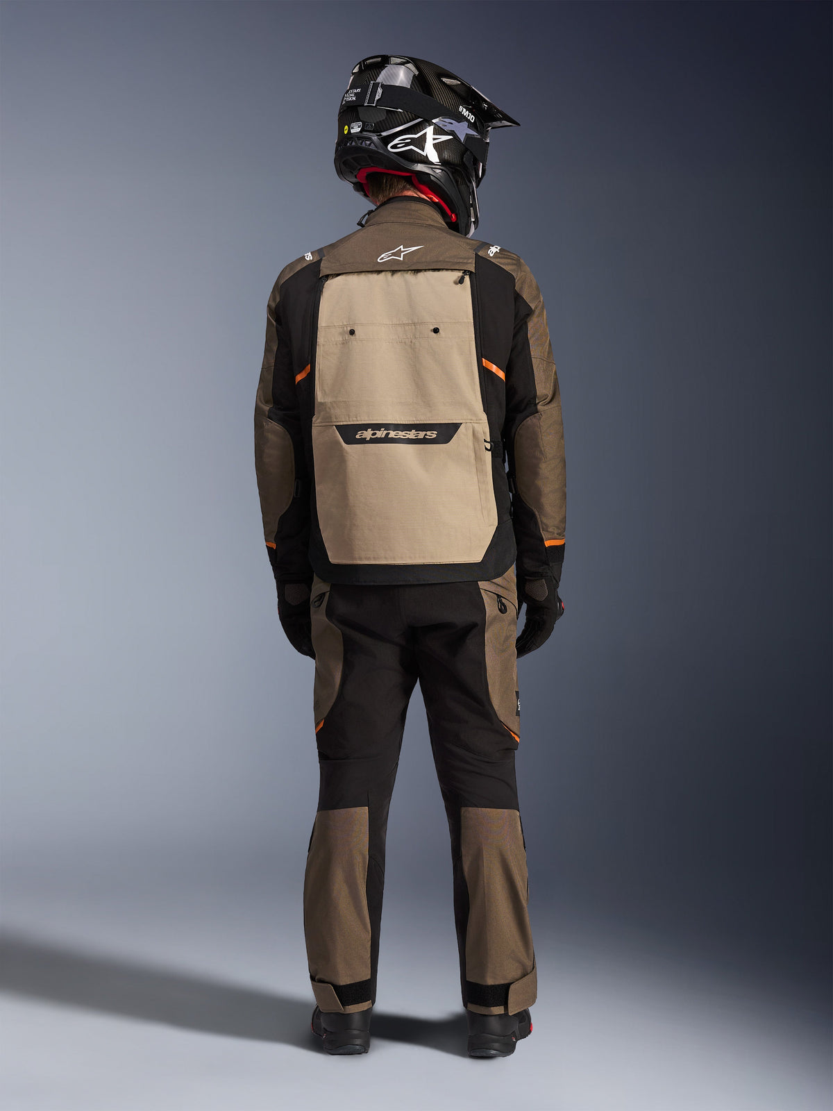Motorcyklist set bagfra iført Alpinestars Cusco Drystar® jakke i Walnut Aluminum Black, med stor aluminiumsfarvet ryglomme og orange detaljer, matchende adventure-bukser, sort off-road-hjelm og handsker, slidstærkt tekstil-motorcykeludstyr til touring.