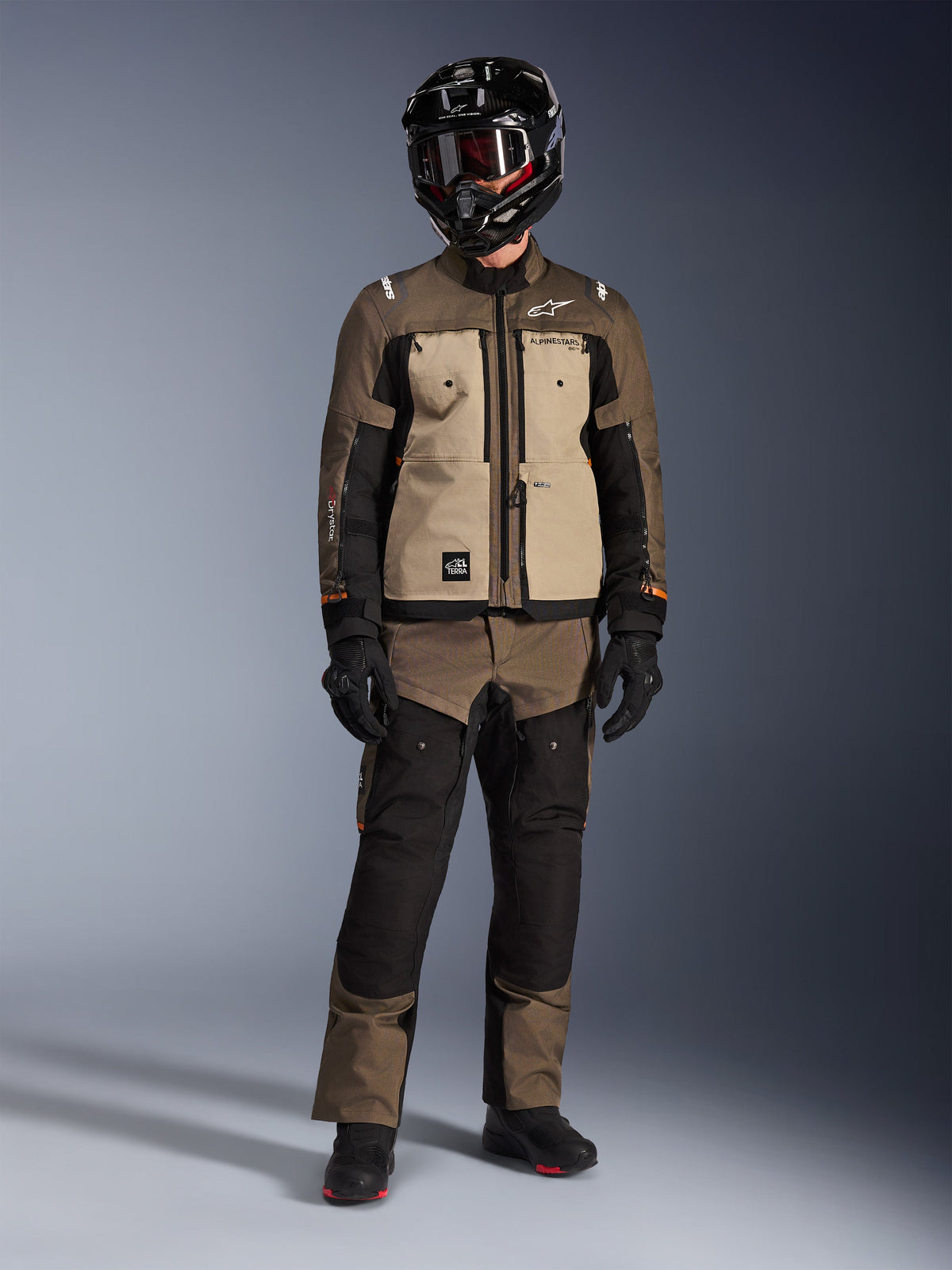 Alpinestars Cusco Drystar® jakke, Walnut Aluminum Black, adventure touring motorcykeljakke båret med matchende bukser og en sort hjelm, med sandfarvede brystpaneler og brune ripstop-ærmer, teknisk køreudstyr til alle vejrforhold