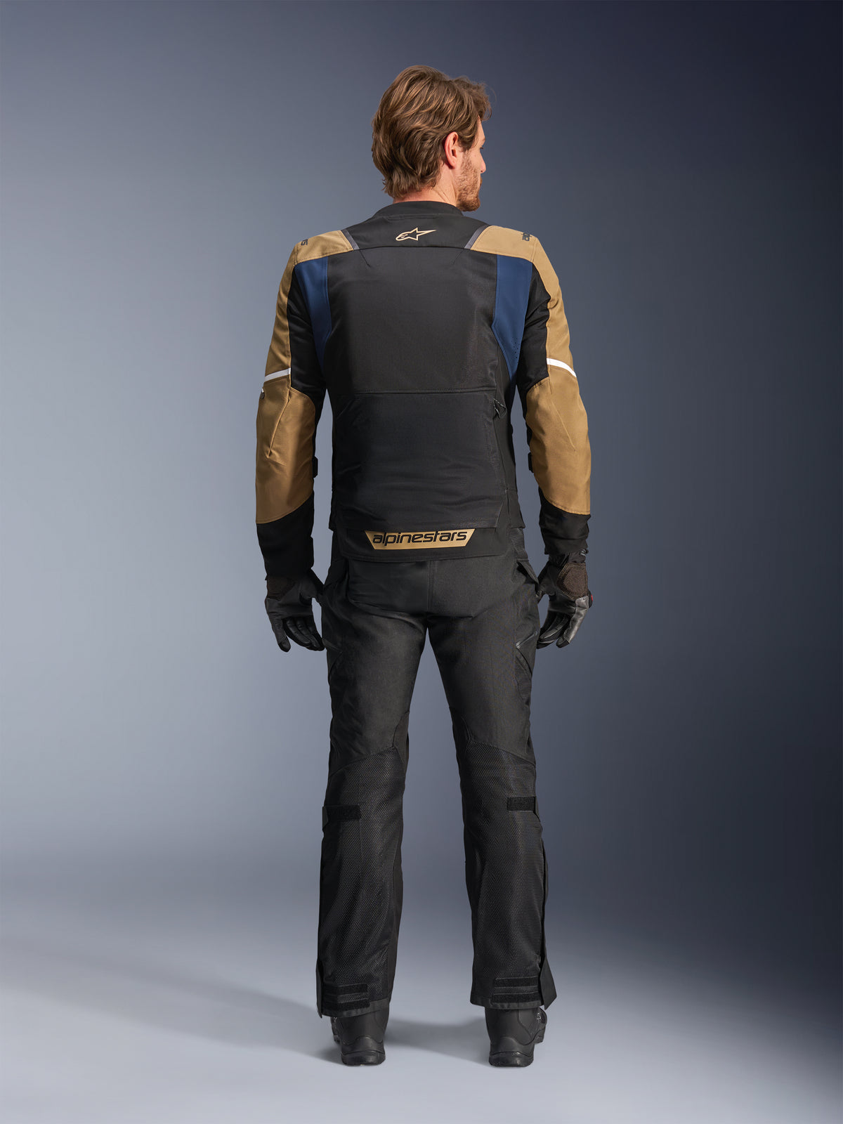 Alpinestars ST-2 Air-jakke i Deep Blue, Dark Khaki og sort, set bagfra på model, der viser det store mesh-panel på ryggen og khaki-farvede ærmer med hvide striber, kombineret med sorte tekniske motorcykelbukser og handsker til adventure touring.