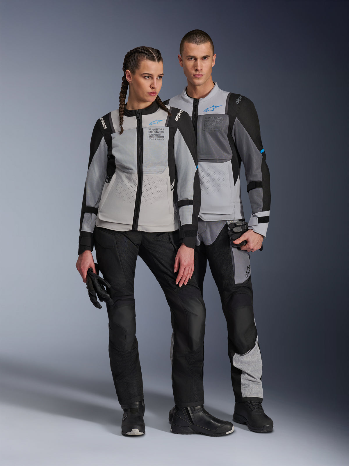 Mandlige og kvindelige modeller iført Alpinestars ST-2 Air-jakker i Ice Gray, Dark Gray og Black, teknisk motorcykeludstyr til sommertouring med store mesh-paneler og exploration equipment-branding, kombineret med sorte kørebukser, handsker og støvler