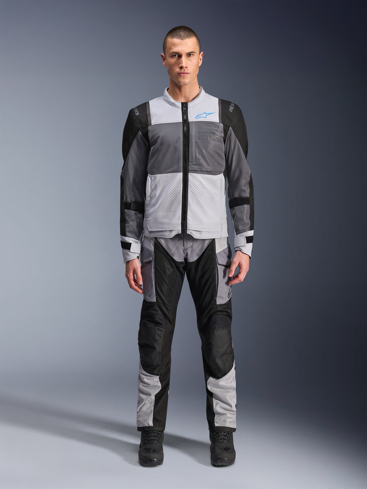 Alpinestars ST-2 Air Jakke, motorcykel touring-jakke, Ice Gray, Dark Gray og sort, båret af en model med matchende adventure-bukser, med store mesh-paneler, blåt Alpinestars-logo på brystet og Alpinestars Exploration Equipment-branding