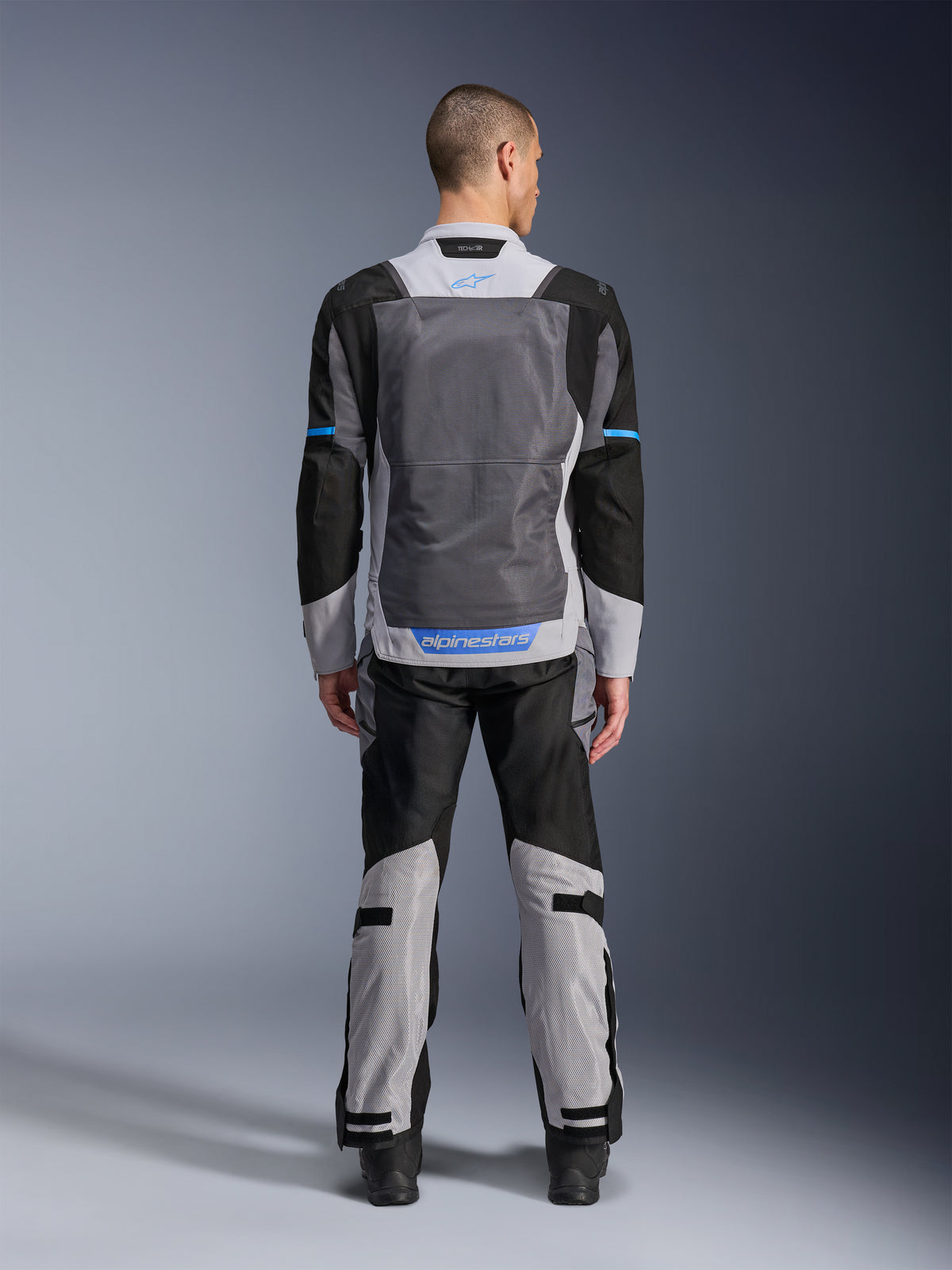 Alpinestars ST-2 Air-jakke, isgrå, mørkegrå og sort, set bagfra på model, med store mesh-ventilationspaneler, blå detaljer og Alpinestars-logo, kombineret med matchende motorcykelbukser til adventure touring.