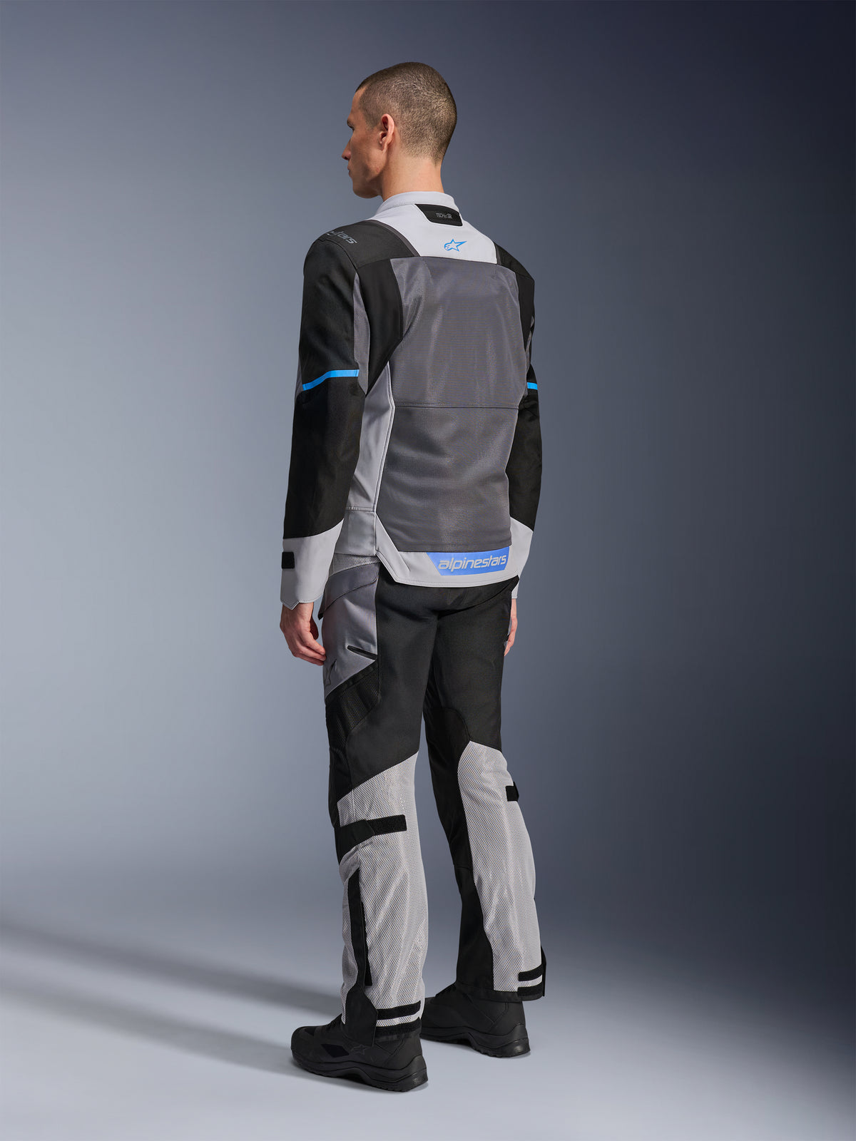 Alpinestars ST-2 Air-jakke, mandlig model set bagfra i Ice Gray, Dark Gray og sort, med stort gråt mesh-rygpanel, blå logo-detaljer og matchende motorcykelbukser til sommer-touring.