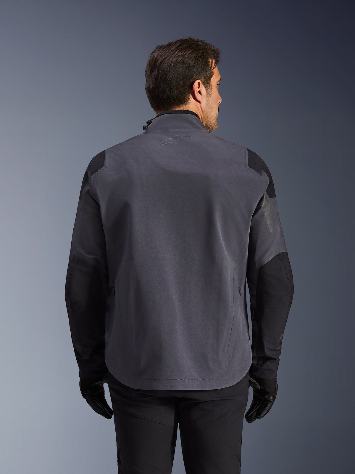 Alpinestars Maxdura Drystar®XF Laminated Armored Shell, teknisk adventure-jakke, mørkegrå og sort, set bagfra med forstærkede skulder- og albuepaneler, diskret logo på den øverste del af ryggen, båret med sorte handsker og bukser, designet til beskyttelse i al slags vejr
