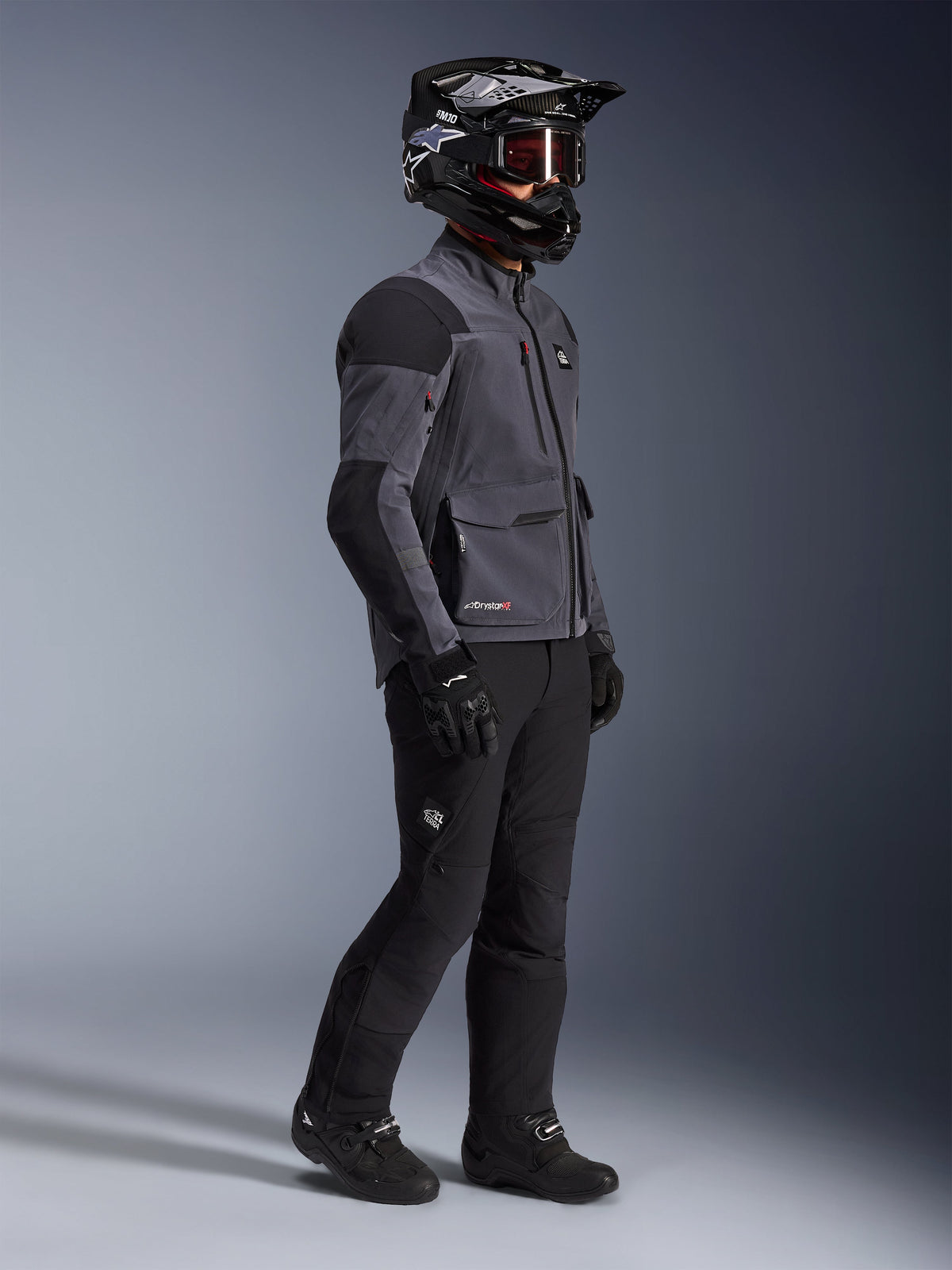 Motorcykelrytter iført Alpinestars Maxdura Drystar®XF Laminated Armored Shell-jakke i mørkegrå og sort, kombineret med sorte adventure-kørebukser, sort hjelm med goggles, sorte handsker og støvler, helkrops-studiebillede der fremhæver det tekniske dual-sport-udstyr.