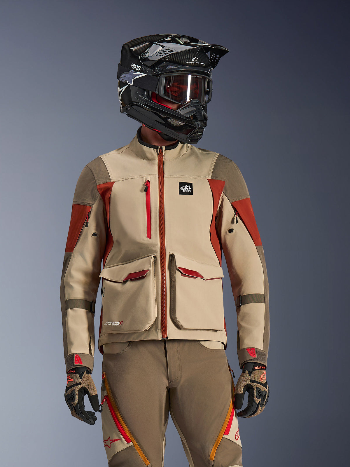 Adventure motorcycle rider wearing the Maxdura Drystar®XF Laminated Armored Shell in Aluminum Walnut Rust Brown, featuring technical 3-layer construction with large front cargo pockets and red accents, paired with a black SM10 helmet, tinted goggles, and matching adventure pants and gloves.