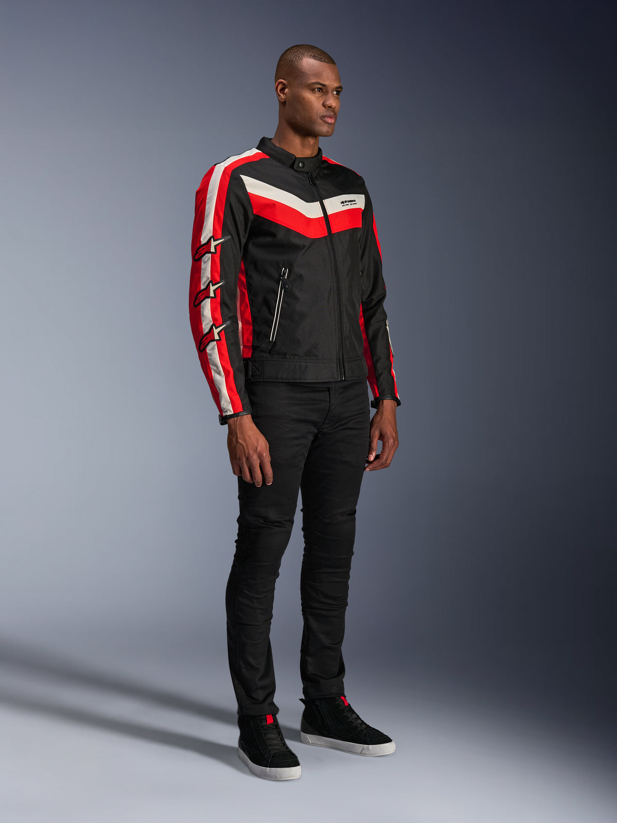 Mandlig model iført Alpinestars T-Dyno WR Jakke, tekstil motorcykeljakke i Black Bright Red Ecru, med røde og råhvide striber og Alpinestars-stjernelogoer på ærmerne, kombineret med sorte bukser og sorte high-top sneakers, vintage-inspireret urbant køreudstyr.