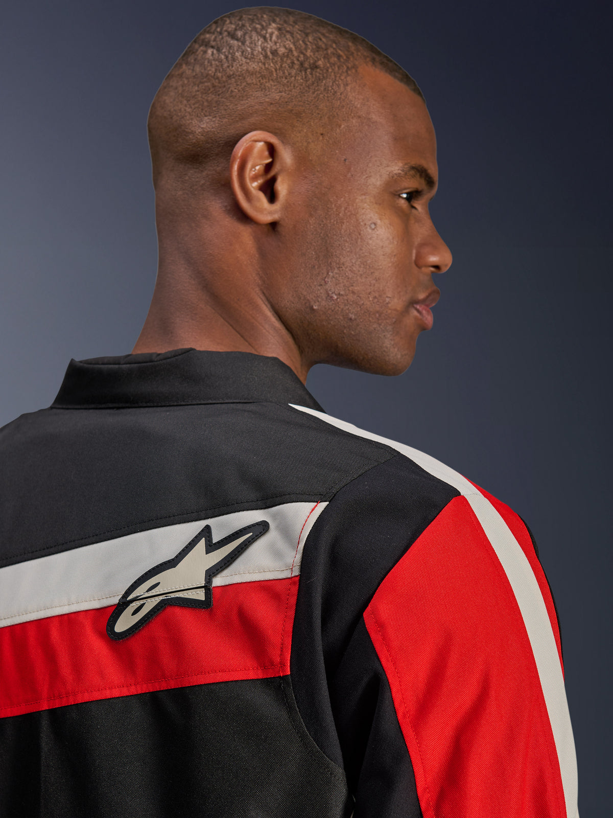 Alpinestars T-Dyno WR-jakke, tæt på-visning bagfra på en model, tekstil-motorcykeljakke i sort med vandrette ecru- og klare røde striber, med Alpinestars stjernelogo-patch på skulderen, vintage-inspireret bikerstil