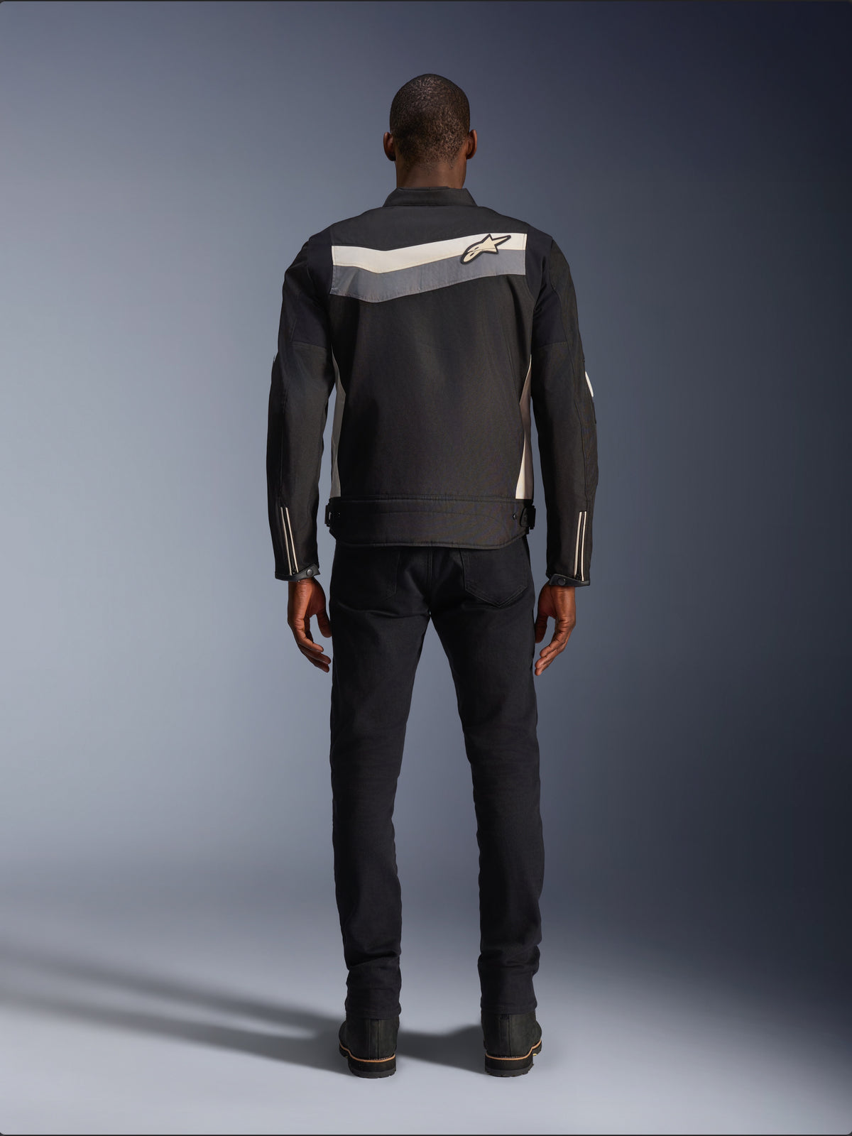 Alpinestars T-Dyno WR-jakke, tekstil motorcykeljakke, sort, mørkegrå og ecru, set bagfra med vandret stribe over skuldrene og lille stjernelogo, båret af model med sorte bukser og støvler, vintage-inspireret køreudstyr