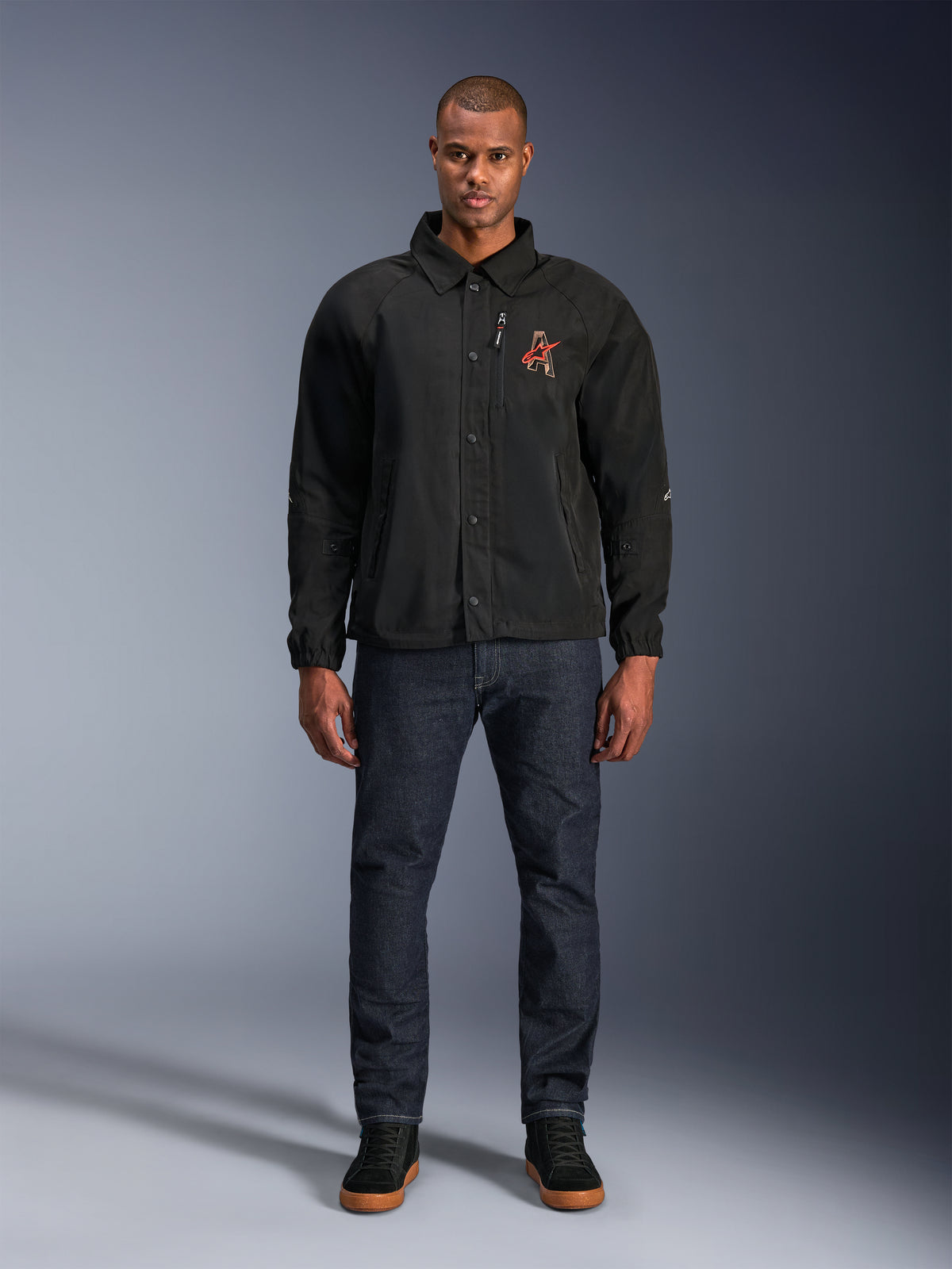 Alpinestars Revved WR-jakke i sort, båret af en model med mørkeblå jeans og sorte sneakers, set forfra, med trykknaplukning og et 3D-broderet logo på brystet, urbant moderne motorcykeludstyr