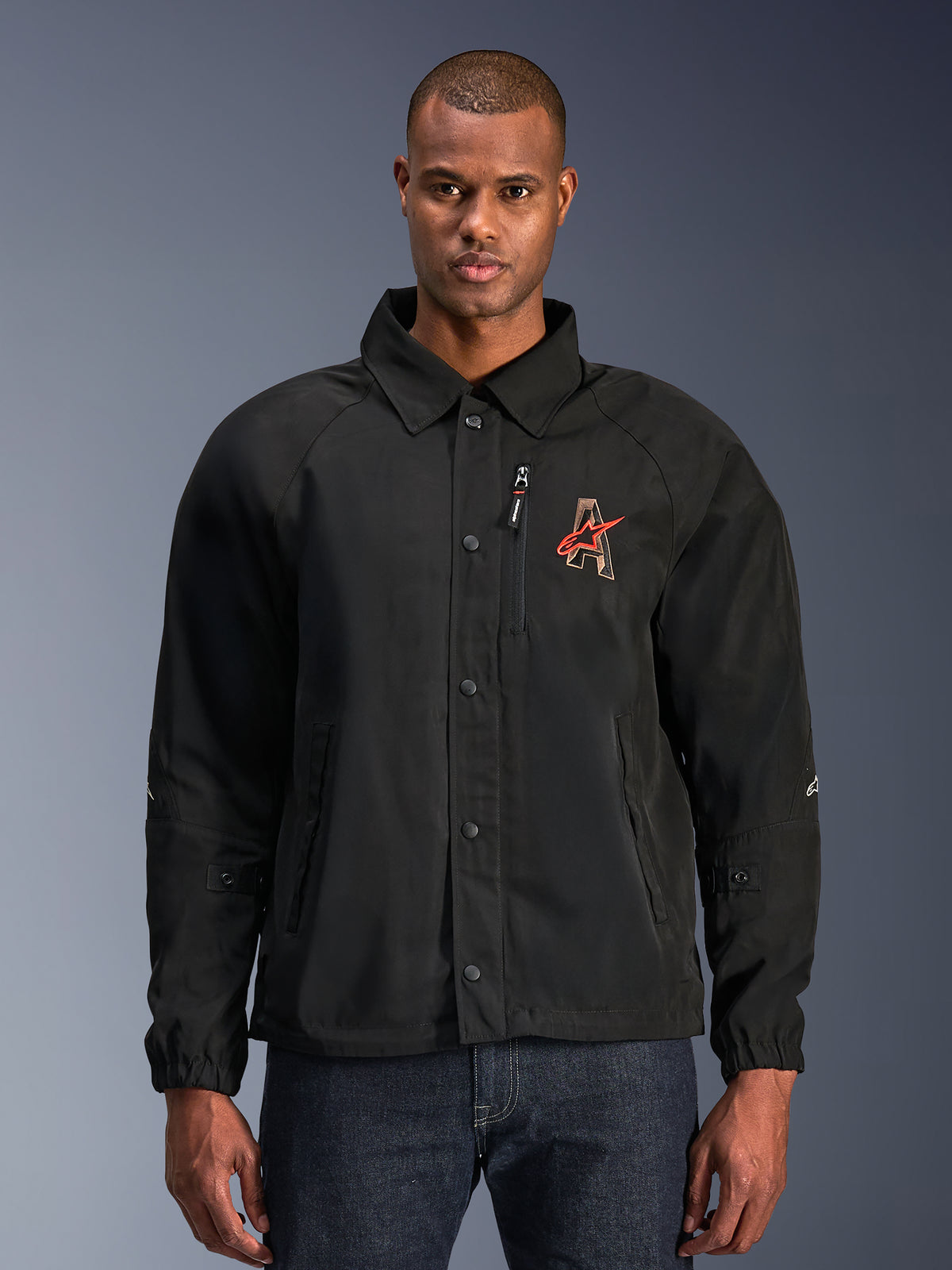 Alpinestars Revved WR-jakke, sort motorcykeljakke med skjortekrave og trykknapper foran, rødt broderet 'A'-logo på brystet, lodret brystlomme med lynlås, elastiske manchetter, urbant coach-jakke design
