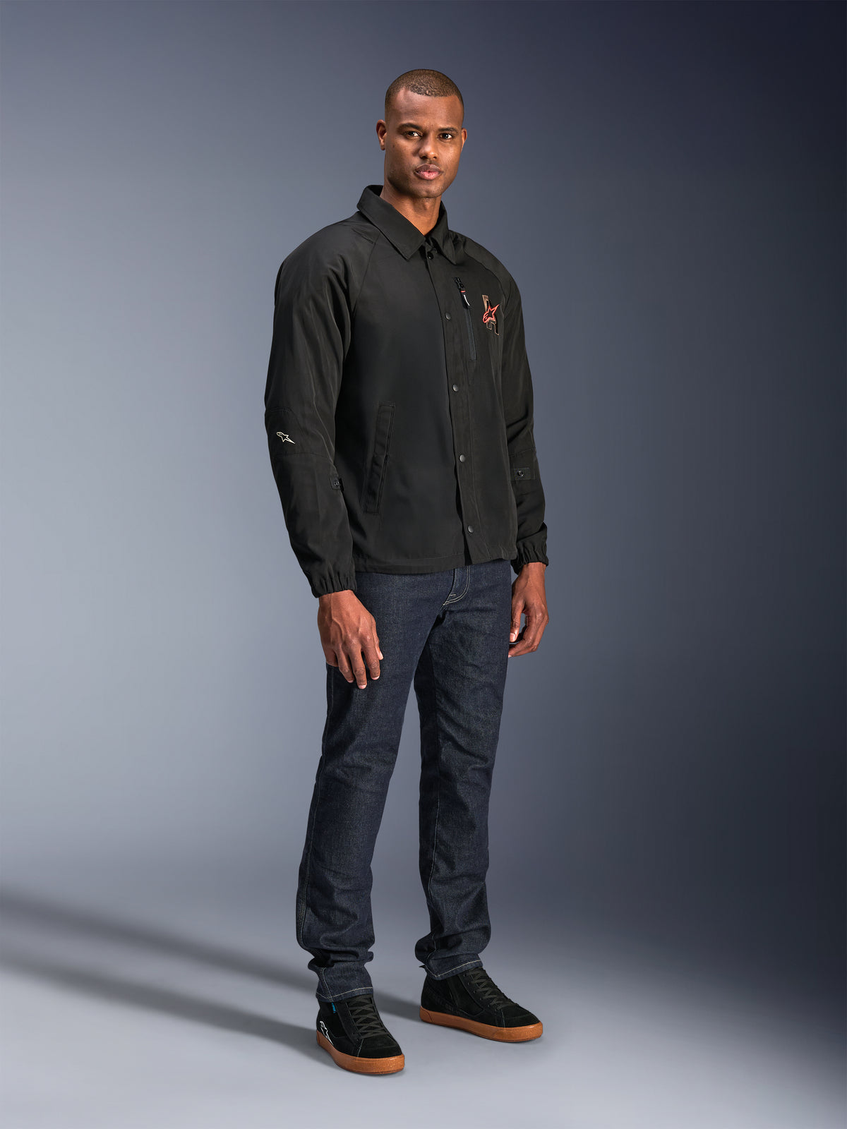 Alpinestars Revved WR-jakke i sort, urban motorcykeljakke i coach-stil, båret af en model med mørkeblå jeans og sorte sneakers, med trykknapper foran og broderet logo, minimalistisk og sporty design til landevejskørsel.