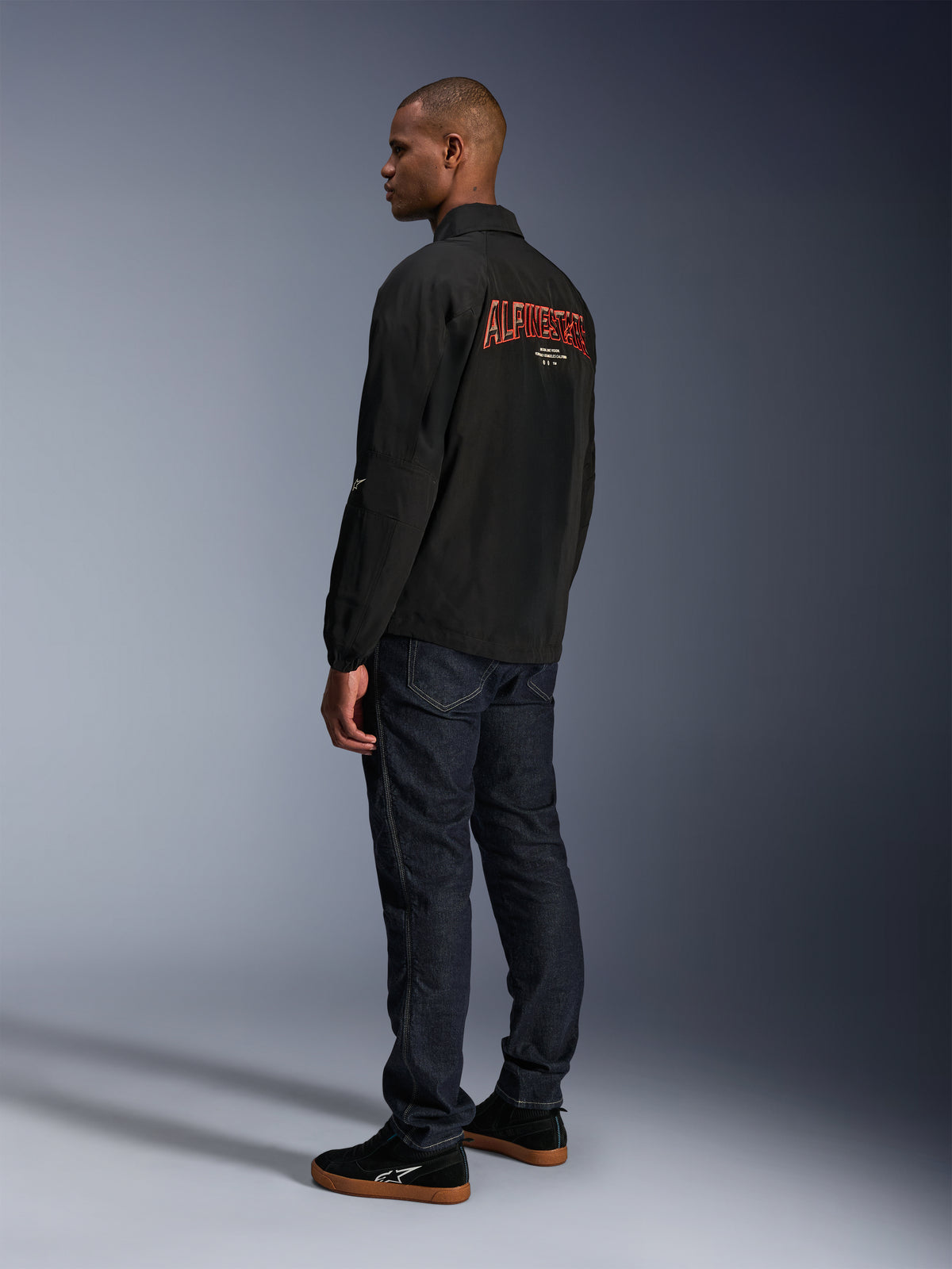 Alpinestars Revved WR-jakke, sort, med stort orange 3D-broderet Alpinestars-branding på ryggen, set bagfra i trekvart profil på en model iført mørkeblå jeans og sorte sneakers, urban moderne motorcykeljakke med coach-jakke-silhuet