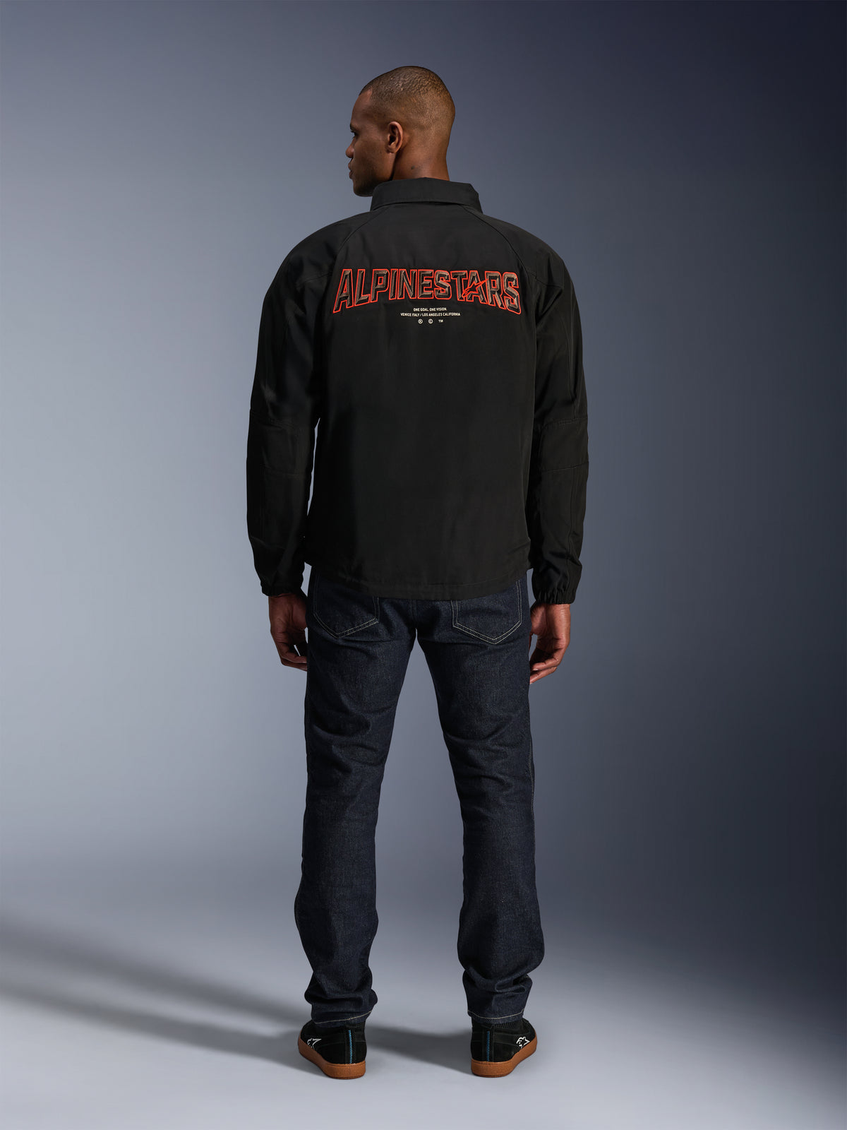 Alpinestars Revved WR Jakke, sort, set bagfra med stort rødt 'ALPINESTARS' broderi i 3D-effekt, urban coach-jakke, båret med mørke denimjeans og sorte sneakers, minimalistisk motorcykelbeklædning
