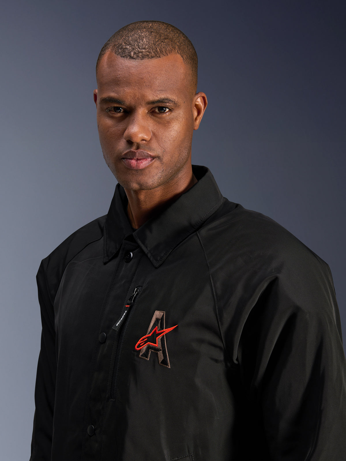 Alpinestars Revved WR Jakke, sort, nærbillede af en mand iført den vandafvisende motorcykeljakke i satin, med 3D-broderet 'A'-logo med rød Alpinestars-stjerne, brystlomme med lynlås og design med krave til moderne bykørsel.