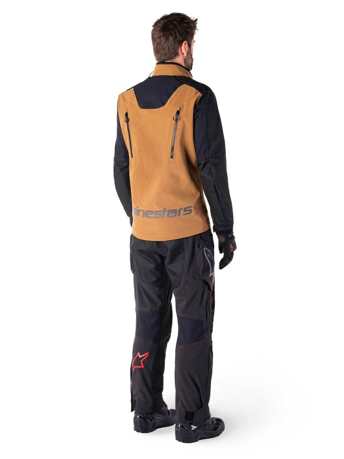 Alpinestars Hyde XT Stretch Drystar®XF Jacket and Hyde XT Drystar® Pants, rear view of a rider wearing tan and black adventure touring gear, jacket featuring vertical back zippers and Alpinestars branding, black pants with red star logo on the calf, paired with black gloves and boots.