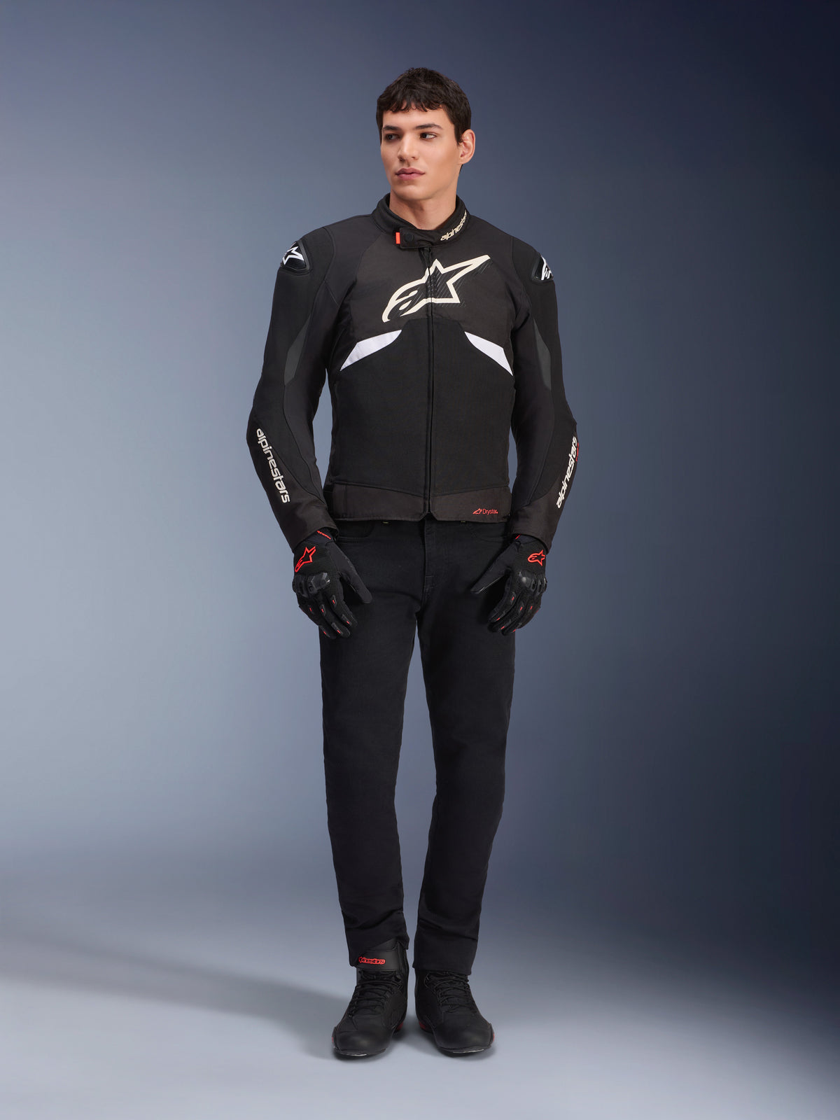 Mandlig model iført Alpinestars T-GP R V3 Drystar®-jakke i sort og hvid, vandtæt sportstekstiljakke med stort hvidt Alpinestars-logo på brystet, kombineret med sorte ridebukser og handsker, professionelt studioportræt, der fremhæver den strømlinede sportsilhuet.