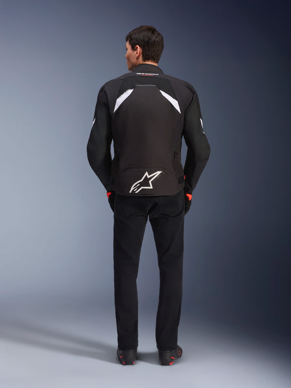 Alpinestars T-GP R V3 Drystar® jakke, sort og hvid, set bagfra, vandtæt tekstil-motorcykeljakke med et stort hvidt stjernelogo nederst på ryggen og hvide detaljer på skuldrene, kombineret med sorte bukser og støvler, race-afledt sportsdesign