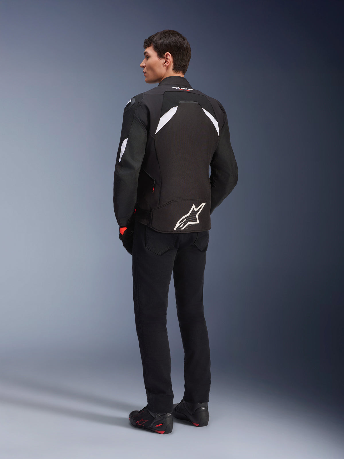 Mandlig model set bagfra iført Alpinestars T-GP R V3 Drystar® jakke i sort og hvid, med hvide detaljer på skuldrene og et stort hvidt logo på lænden, kombineret med sorte mc-bukser og støvler, sportstekstil motorcykeljakke.