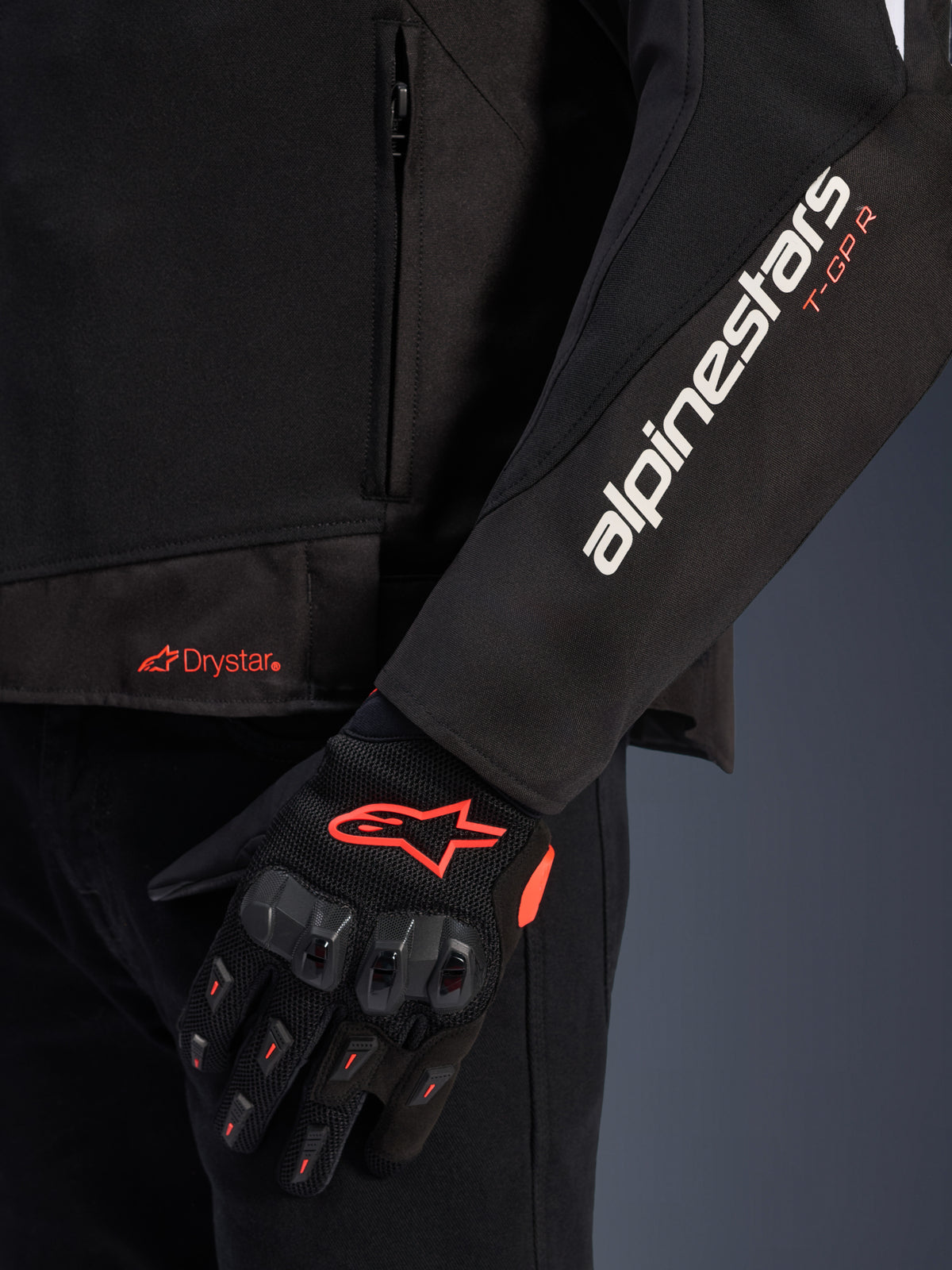 Alpinestars T-GP R V3 Drystar® jakke, sort tekstil motorcykeljakke, nærbillede der viser hvidt Alpinestars og orange T-GP R logo på ærmet, orange Drystar-logo på kanten, kombineret med sorte beskyttende mesh-handsker med røde detaljer.