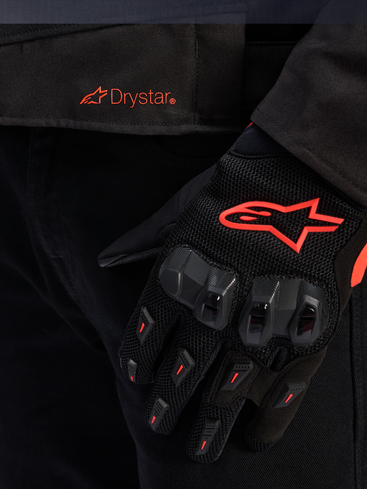 Nærbillede af taljen på Alpinestars T-GP R V3 Drystar® jakken med et rødt Drystar-logo, vist med en sort motorcykelhandske med et rødt Alpinestars stjernelogo, mesh-stof og forstærket knobeskkyttelse.