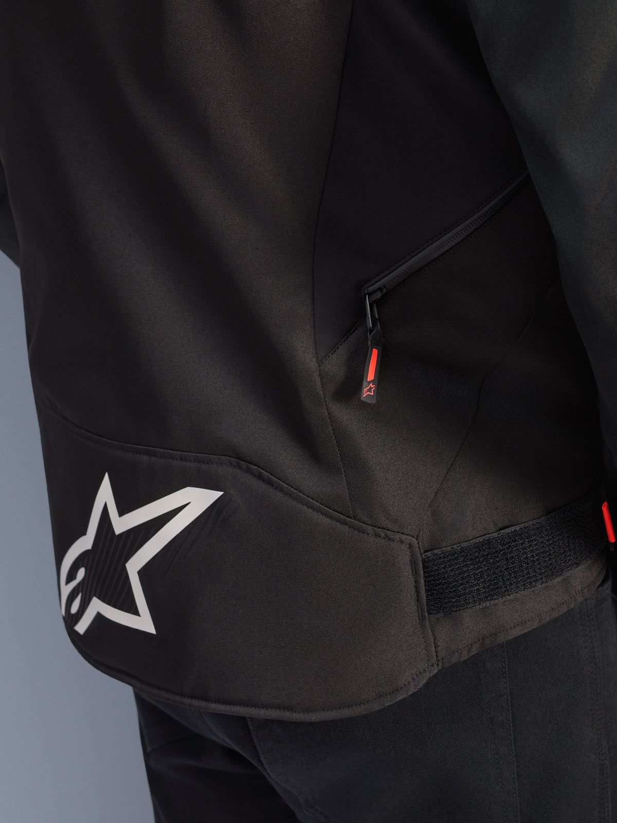 Alpinestars T-GP R V3 Drystar®-jakke, sort og hvid, nærbillede af den nederste rygsektion med et stort hvidt Alpinestars-stjernelogo, sidelomme med lynlås og rød trækstrop, velcro-justeringsstrop i taljen, slidstærkt tekstilmateriale.