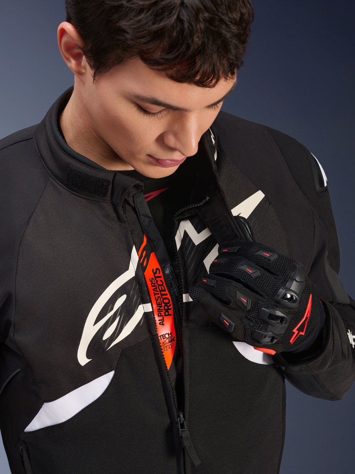 Alpinestars T-GP R V3 Drystar®-jakke, sort og hvid sportstekstiljakke til motorcykel, delvist lynet op så den orange 'Alpinestars Protects' indvendige strop ses, båret med sorte og røde mesh-handsker, race-afledt beskyttelsesbeklædning.