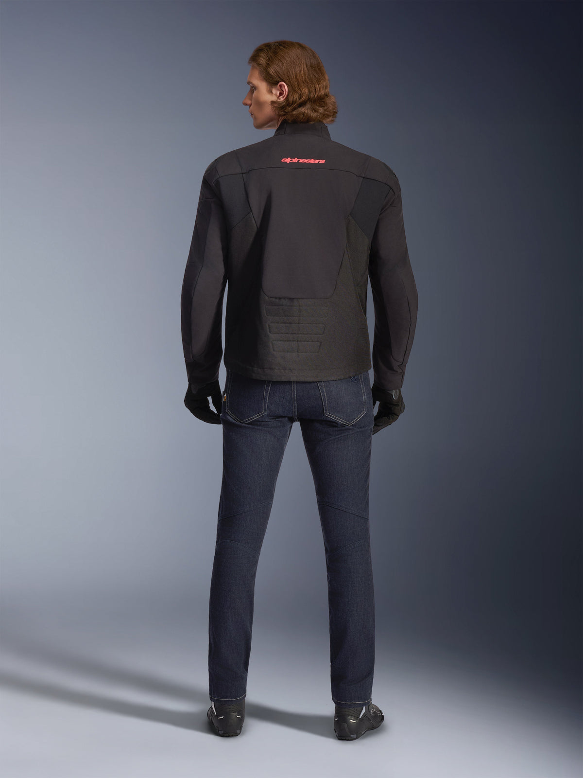 Alpinestars SMX Vandtæt Jakke, sort, set bagfra på en model, med rødt logo på øverste del af ryggen, forlænget rygstykke for bedre dækning under kørsel, stylet med mørkeblå motorcykeljeans og sorte handsker.