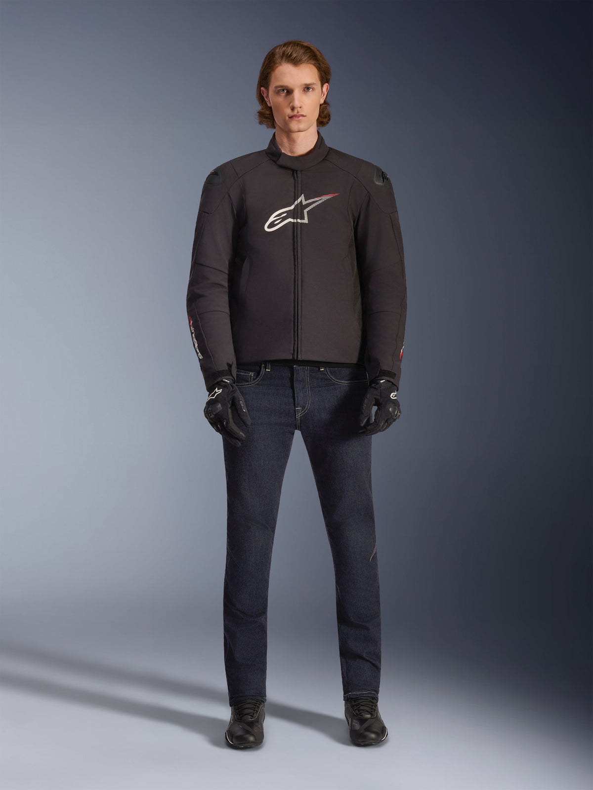 Model iført Alpinestars SMX Vandtæt Jakke i sort med hvidt og rødt logo, kombineret med mørke denim motorcykelbukser, sorte handsker og motorcykelstøvler, sporty motorcykeludstyr til al slags vejr