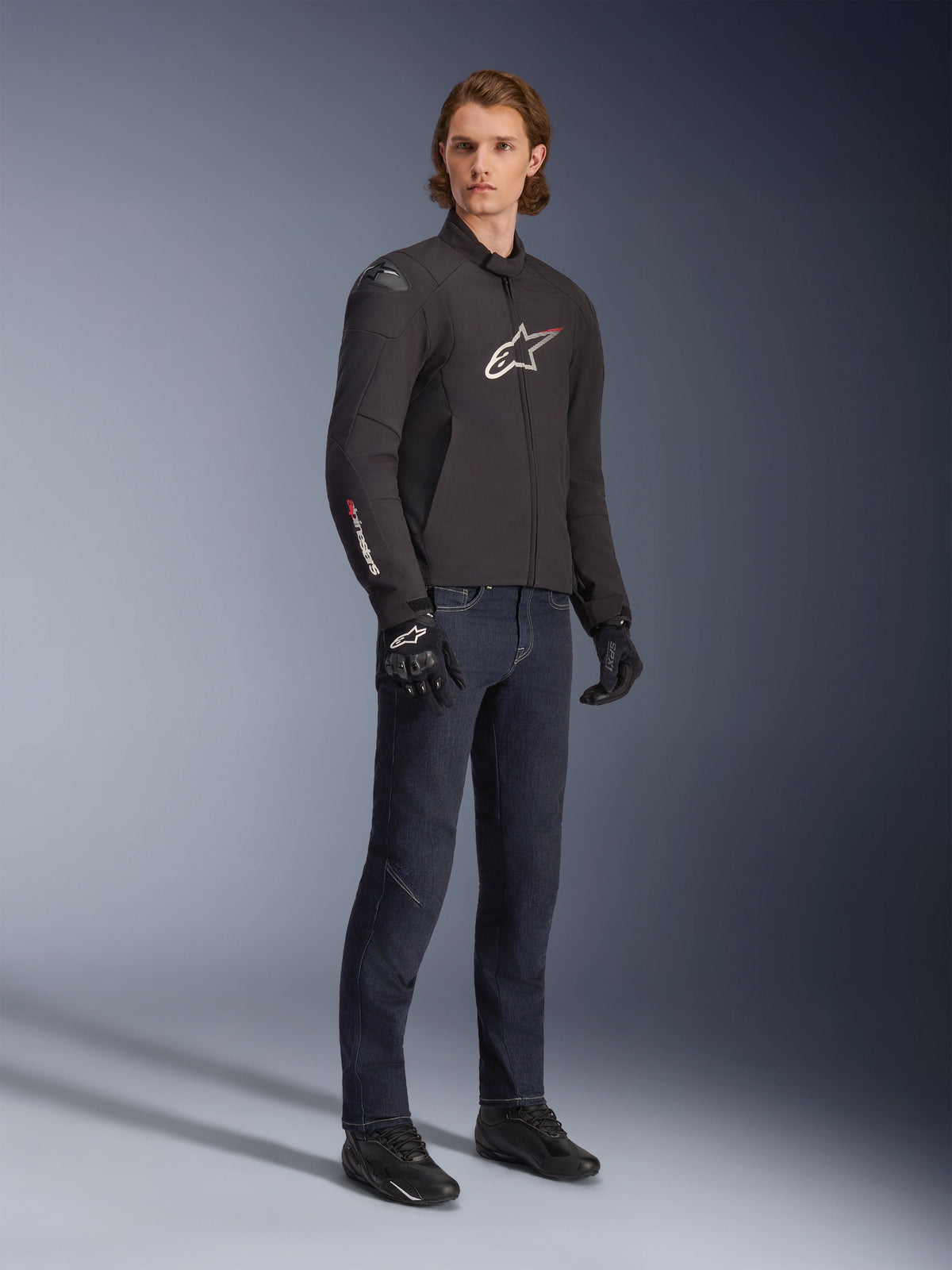 Mandlig model iført Alpinestars SMX vandtæt jakke, sort med hvide og røde logo-detaljer, stylet med mørkeblå denimbukser og sorte kørehandsker, strømlinet sport-fit tekstil-motorcykeljakke