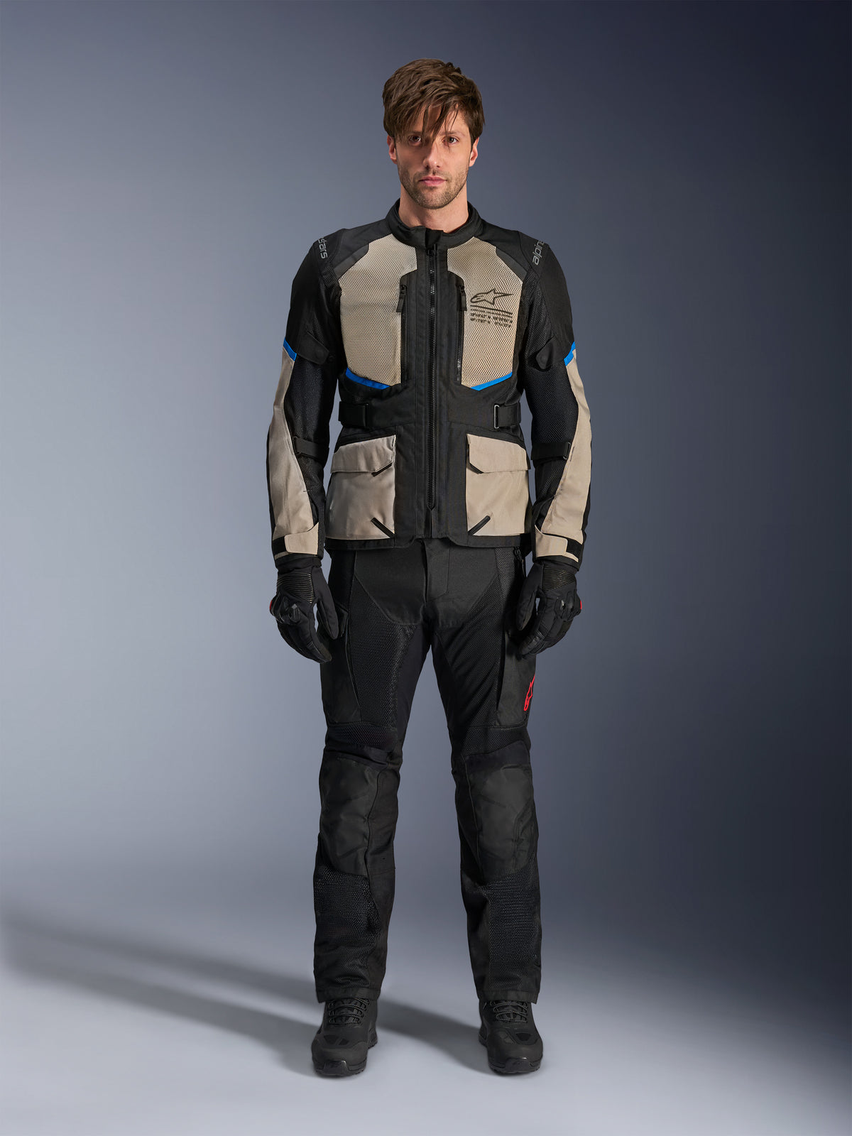 Model iført Alpinestars Andes Air Drystar® jakke, motorcykel touring-jakke, sort, Laurel Gray og lyseblå, kombineret med matchende sorte kørebukser og handsker, med omfattende mesh-ventilationspaneler på brystet og ærmerne til adventure touring i varmt vejr.