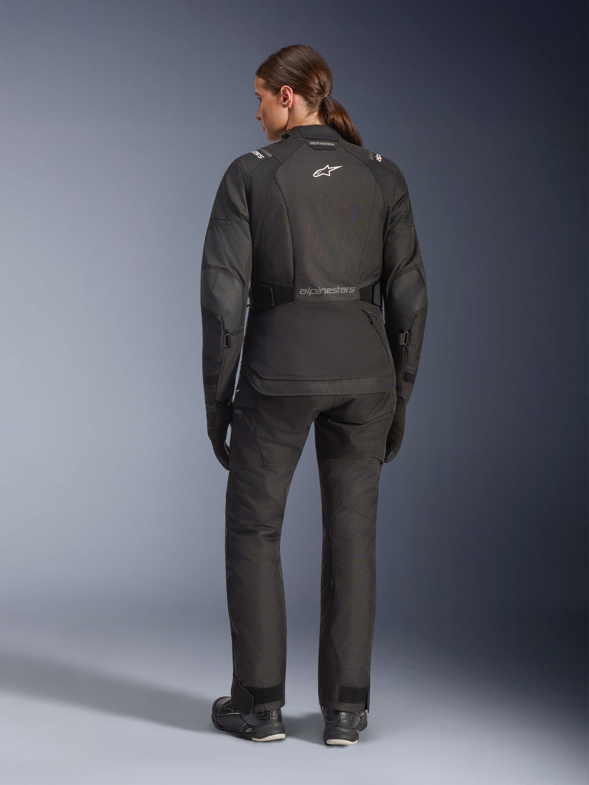 Kvinde set bagfra iført Alpinestars Stella Andes V4 Drystar® jakke, sort, matchende motorcykelbukser, hvidt Astar-logo på ryggen, forlænget rygstykke, komplet adventure-touring køreudstyr inklusiv sorte handsker og støvler