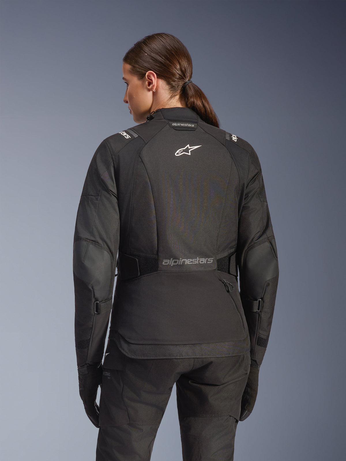 Kvinde iført Alpinestars Stella Andes V4 Drystar-jakke i sort, set bagfra med hvidt logo på ryggen og forstærkede albuepaneler, kombineret med matchende sorte bukser og handsker, adventure touring motorcykelsæt