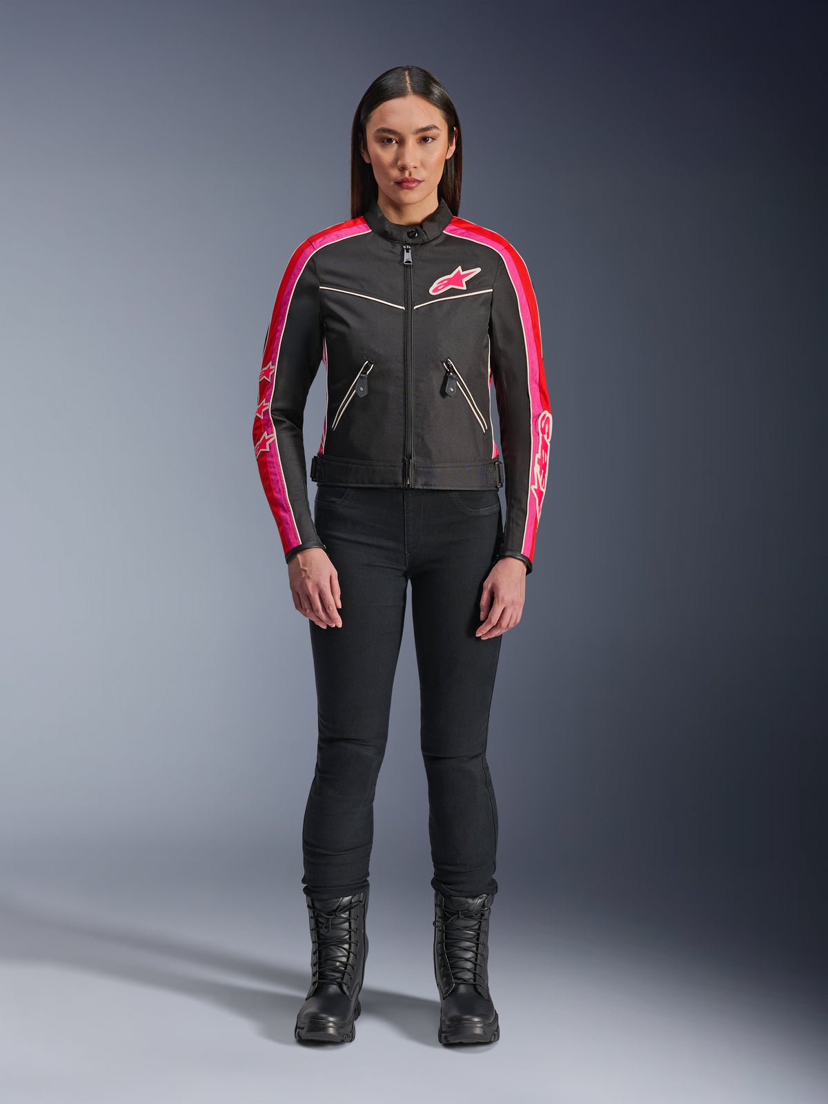 Kvinde iført Alpinestars Stella T-Dyno WR-jakke, sort med klare røde og fuchsia-farvede detaljer, tekstiljakke i vintage-stil med lyserødt logo og striber på ærmerne, kombineret med sorte bukser og snørestøvler, urbant motorcykeludstyr