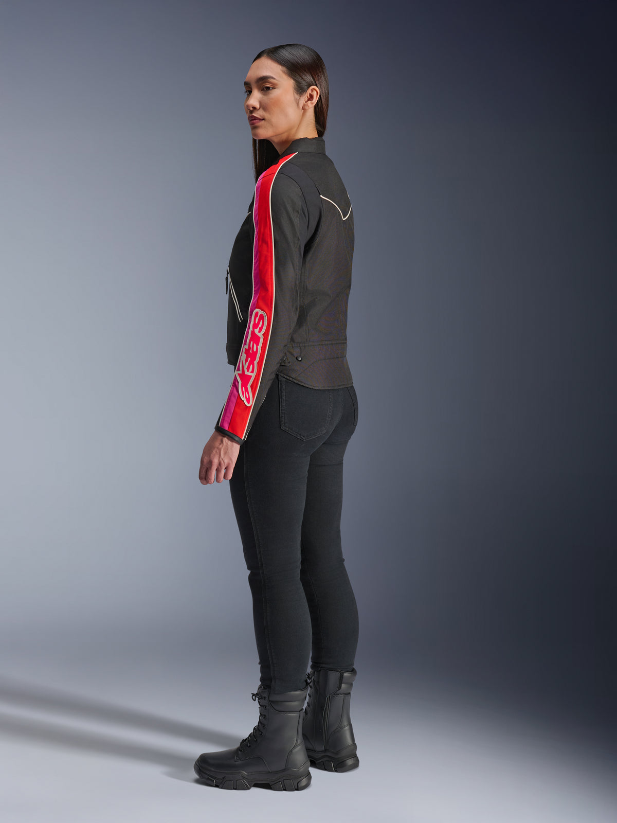 Kvindelig model iført Alpinestars Stella T-Dyno WR-jakke i Black Bright Red Fuchsia, med markante røde og lyserøde striber og Alpinestars-branding på ærmet, kombineret med sorte motorcykelbukser og sorte motorcykelstøvler i et studiemiljø.