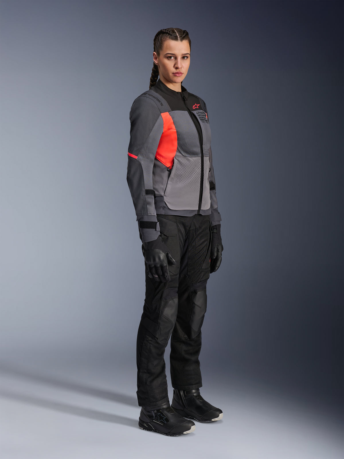 Kvindelig motorcykelrytter iført Alpinestars Stella ST-2 Air-jakke i Deep Gray, Dark Gray og Fluo Coral, kombineret med sorte motorcykelbukser, handsker og støvler, moderne touring-design med mesh-paneler og reflekterende detaljer.