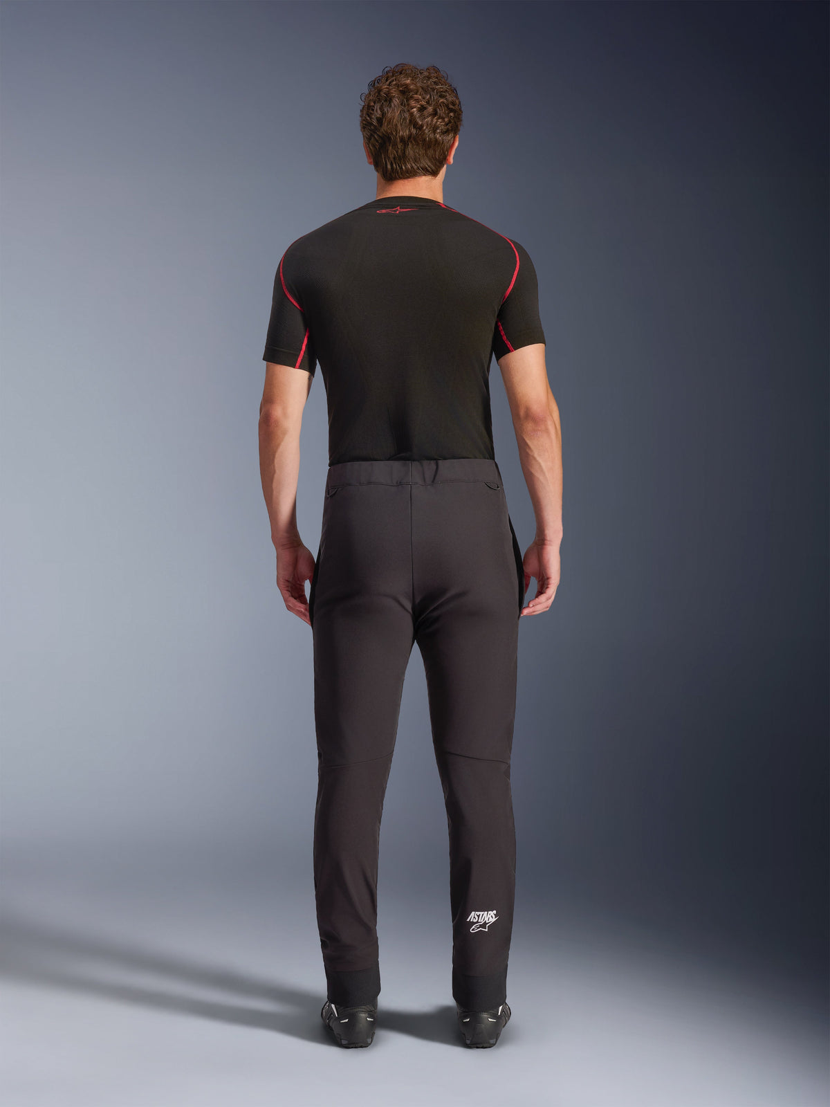 Alpinestars Aeroshell bukser, crossover sports-softshell motorcykelbukser, sort, set bagfra, vist med et sort kortærmet basislag, tætsiddende pasform, forbuede knæ og hvidt Astars-logo på nederste venstre ben.