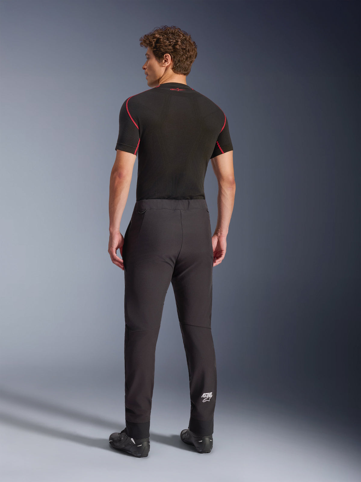 Alpinestars Aeroshell bukser, sort, softshell motorcykelbukser, set bagfra på en model, med en moderne tilspidset pasform, hvidt Astars-logo på underbenet og elastiske kanter, båret med et sort kortærmet baselayer og køresko.