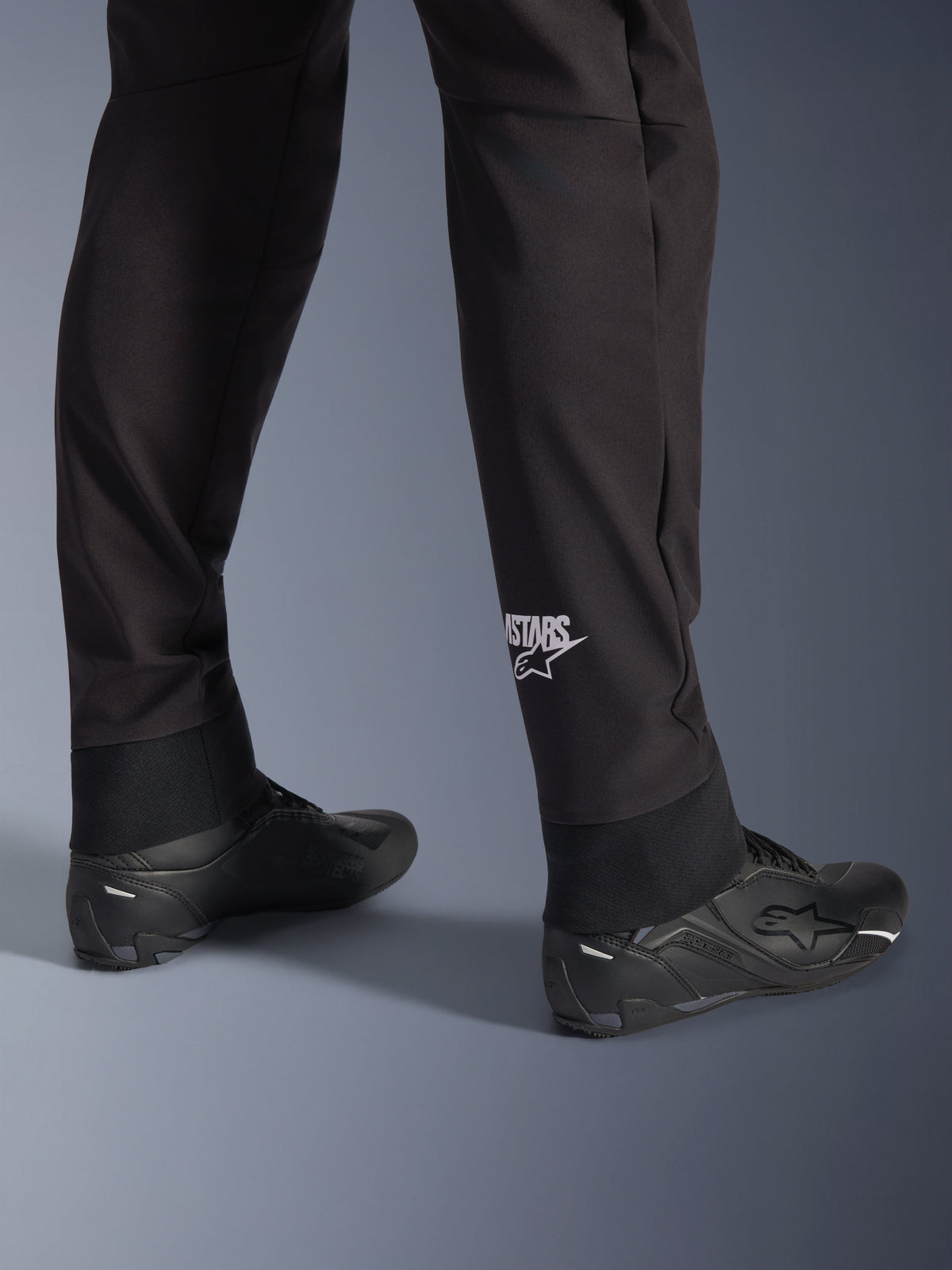 Alpinestars Aeroshell bukser, sorte softshell motorcykelbukser, nærbillede af underben og elastisk kant, hvidt Alpinestars-logo på læggen, stylet med sorte køresko, moderne crossover sportsdesign