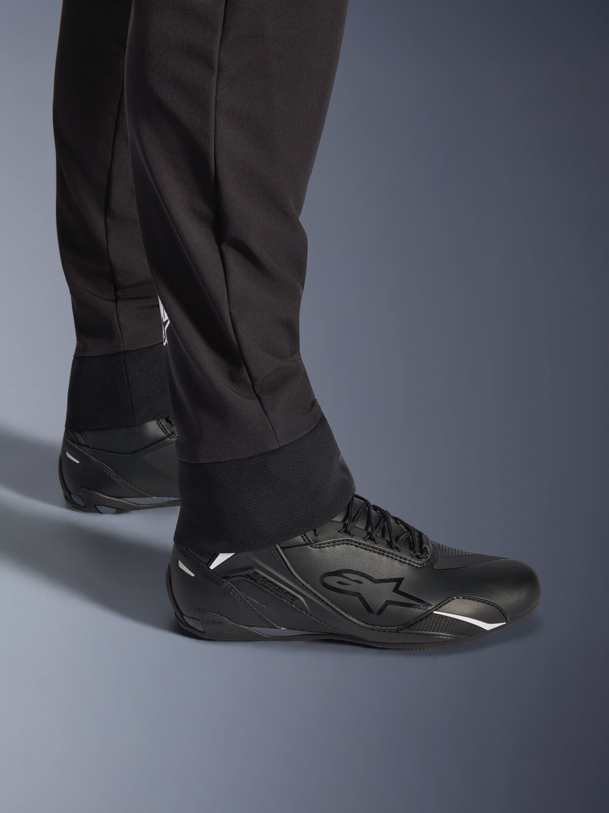 Alpinestars Aeroshell bukser, sort, nærbillede af den elastiske nederste kant og manchet, moderne sports-softshelldesign, båret med sorte Alpinestars køresko.