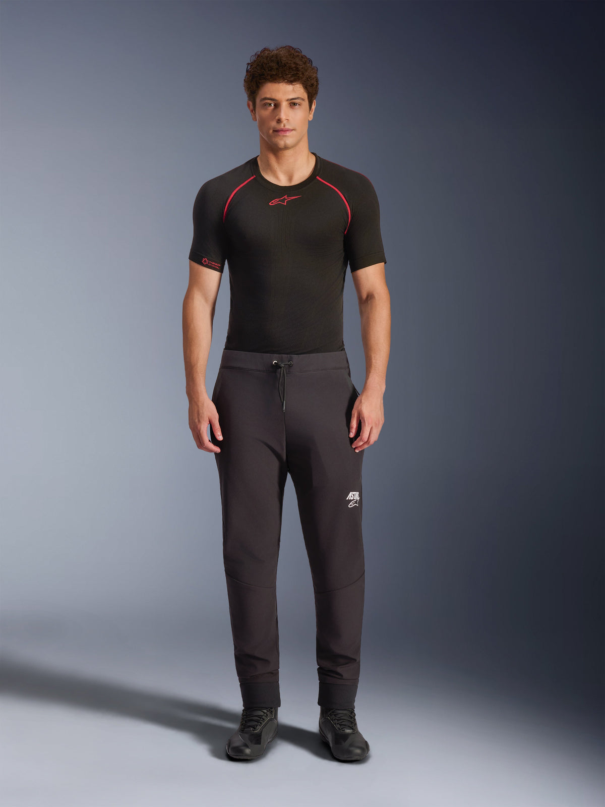 Alpinestars Aeroshell bukser, sorte motorcykel softshell-bukser, vist med et sort kortærmet basislag og sorte sko, med elastisk talje med snørelukning, hvidt Astars-logo på låret og elastiske opslag, moderne atletisk silhuet til landevejskørsel.