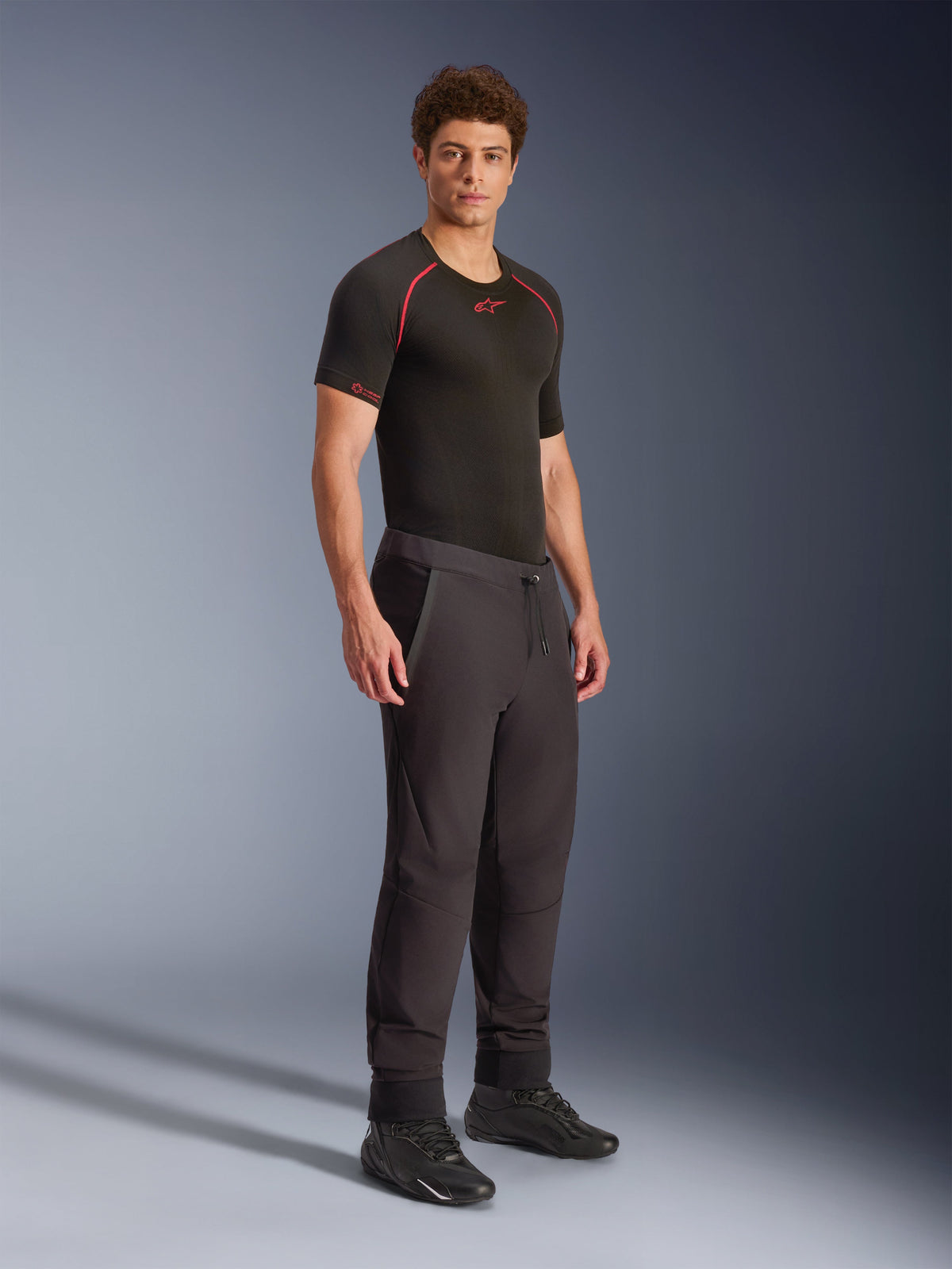Alpinestars Aeroshell-bukser, sorte, crossover sports softshell-motorcykelbukser, båret af en mandlig model med sort performance-basislag og sorte sko, med snøre i taljen og elastiske kanter.